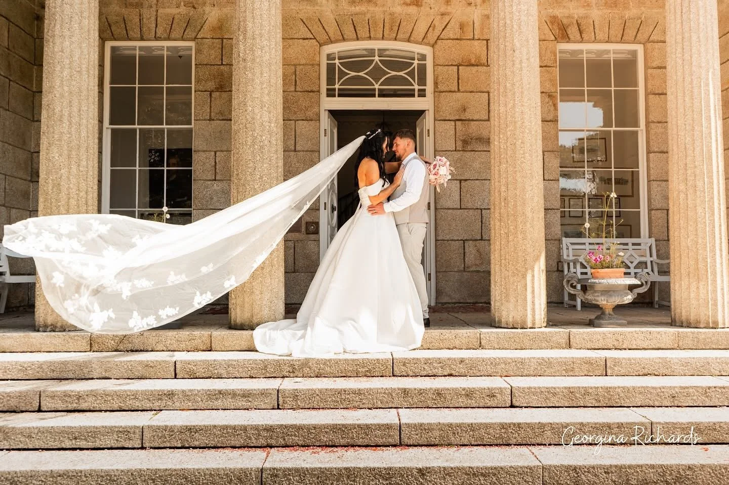 A bride and groom standing on the steps in front of a large stone building with columns, sharing a romantic moment and looking into each other's eyes. The bride's long veil flows behind her.