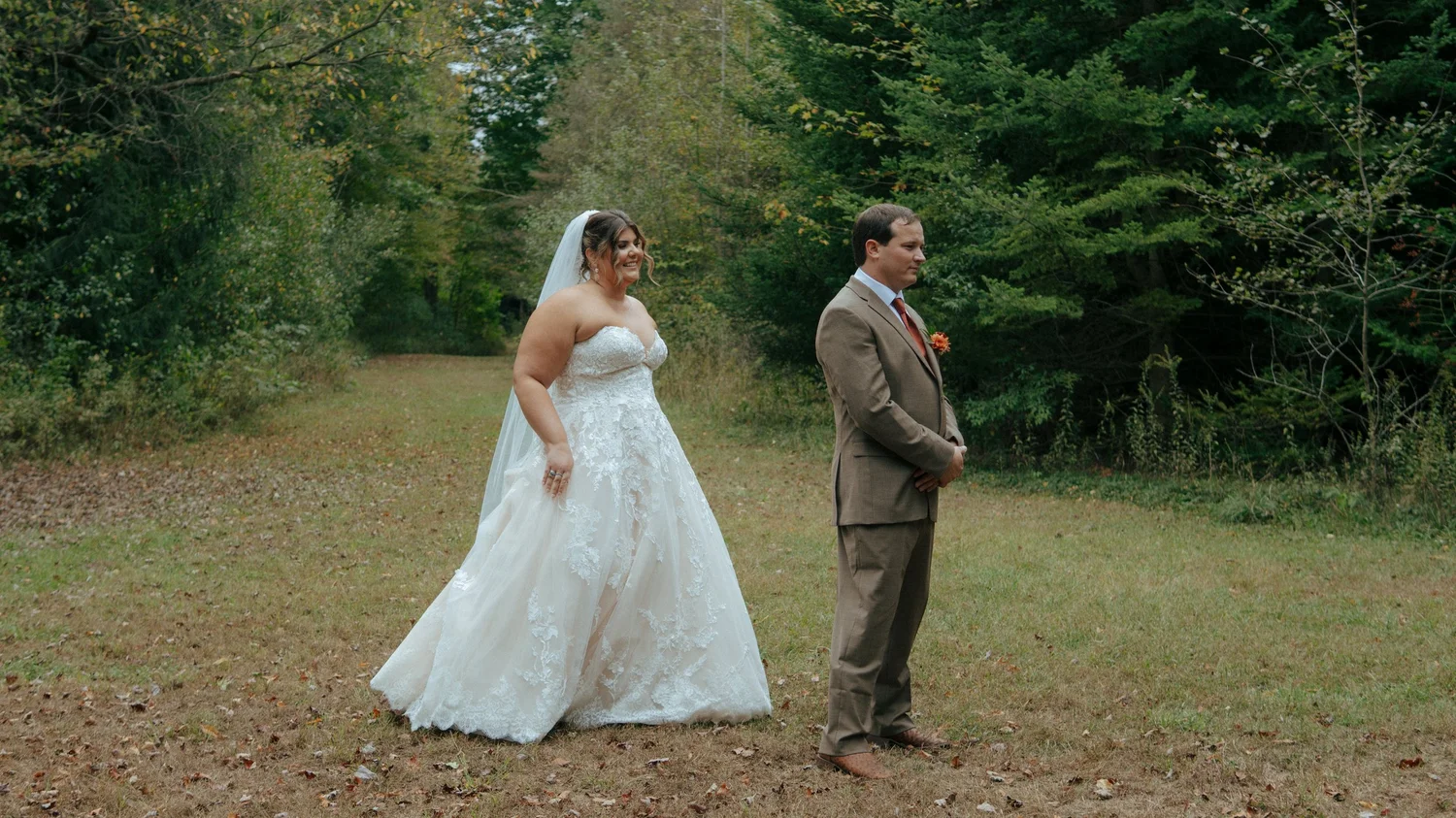A bride and groom walking through a green field, surrounded by trees.