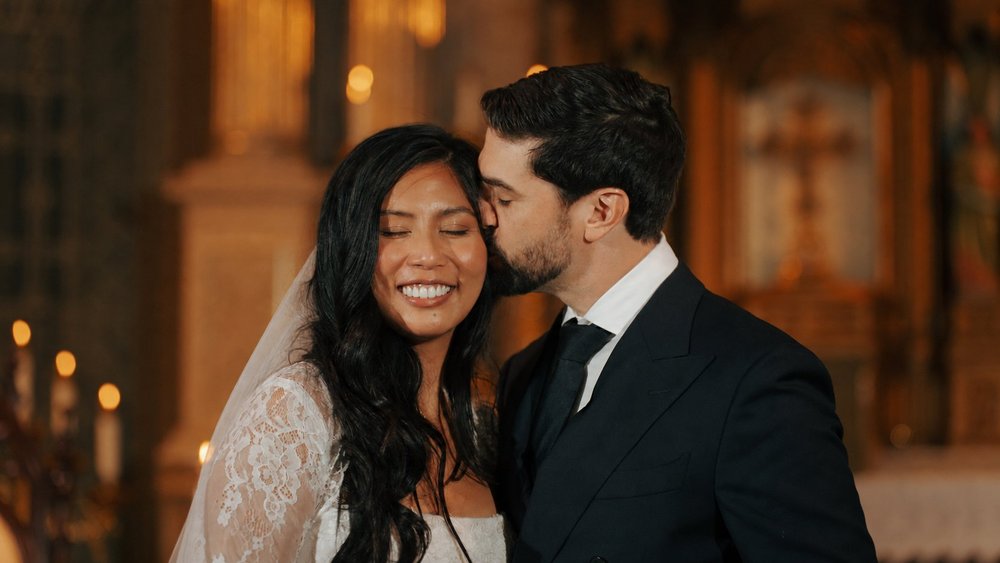 A smiling bride leans toward her partner in a warmly lit room.
