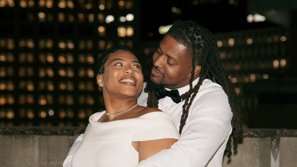 A smiling couple embracing outdoors at night, with lit windows behind them.