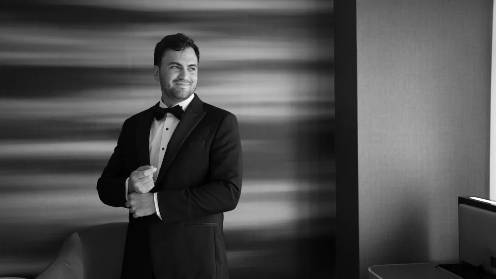 Black-and-white portrait of a groom standing beside a softly lit wall.