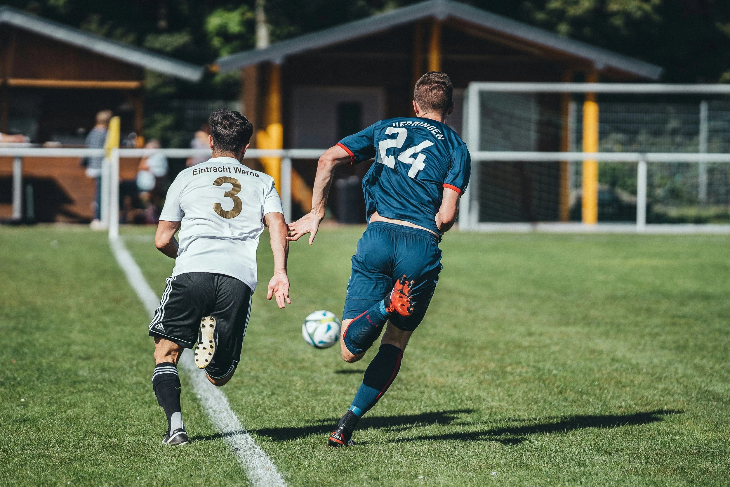 Two soccer players running on a grass field during a game, with a goalpost and trees in the background.