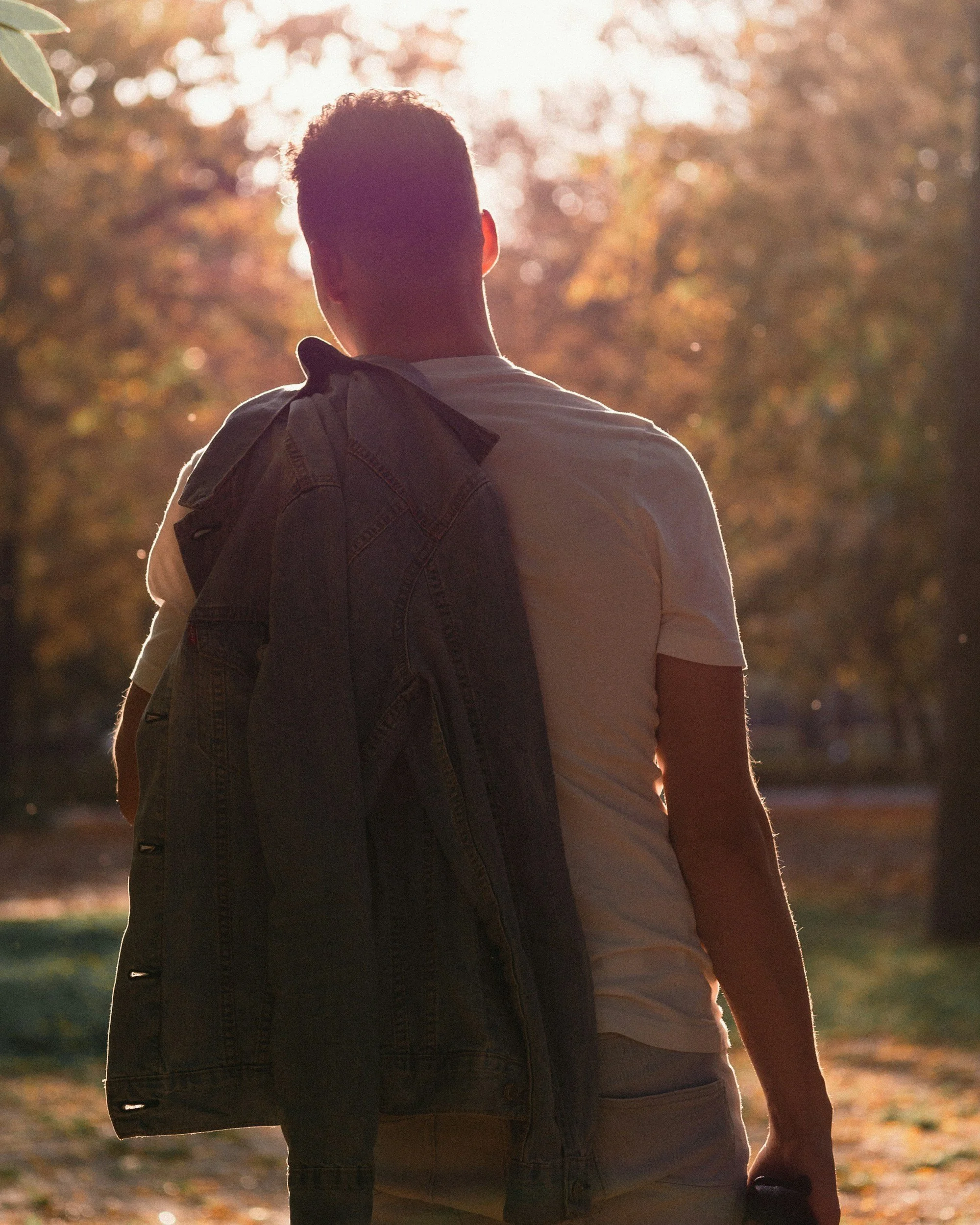 A man and a woman walking outdoors at sunset, seen from behind. The man is carrying a denim jacket over his shoulder and the woman is wearing a short-sleeved shirt. The background shows blurred trees with autumn-colored leaves.