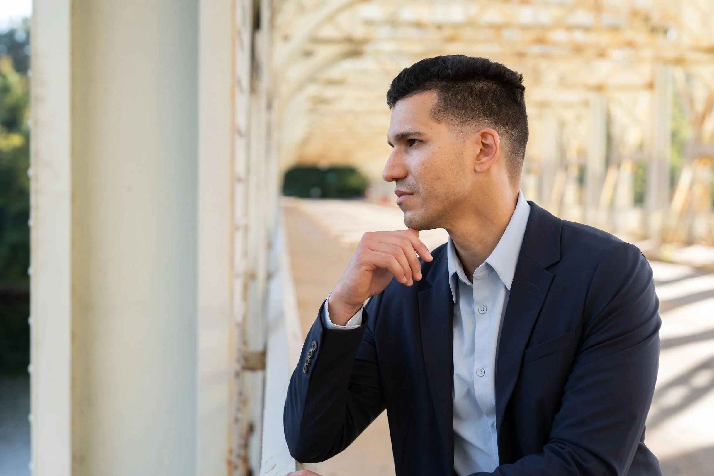 A man in a suit sitting outdoors near a white wooden structure, with his chin resting on his hand, looking thoughtful.