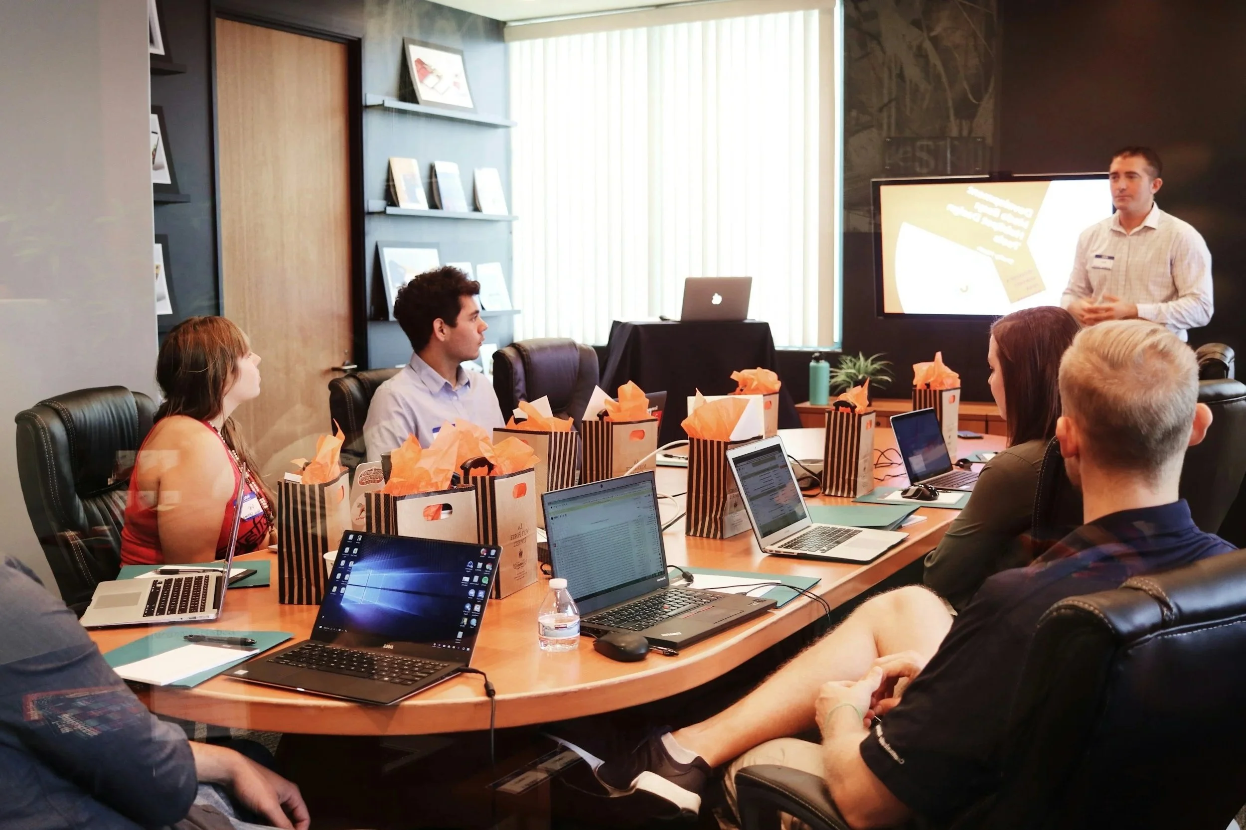 A group of six people attending a business meeting in a conference room, with one person standing and speaking at a presentation screen, while others are seated around a large table with laptops and gift bags.