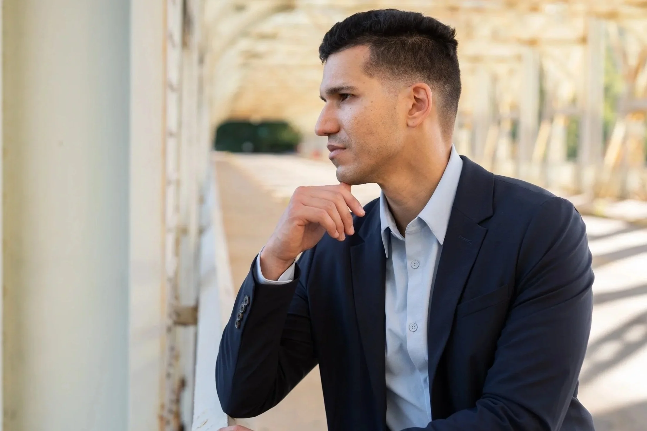 A man in a dark suit and white shirt sitting outdoors, resting his chin on his hand, looking thoughtful.
