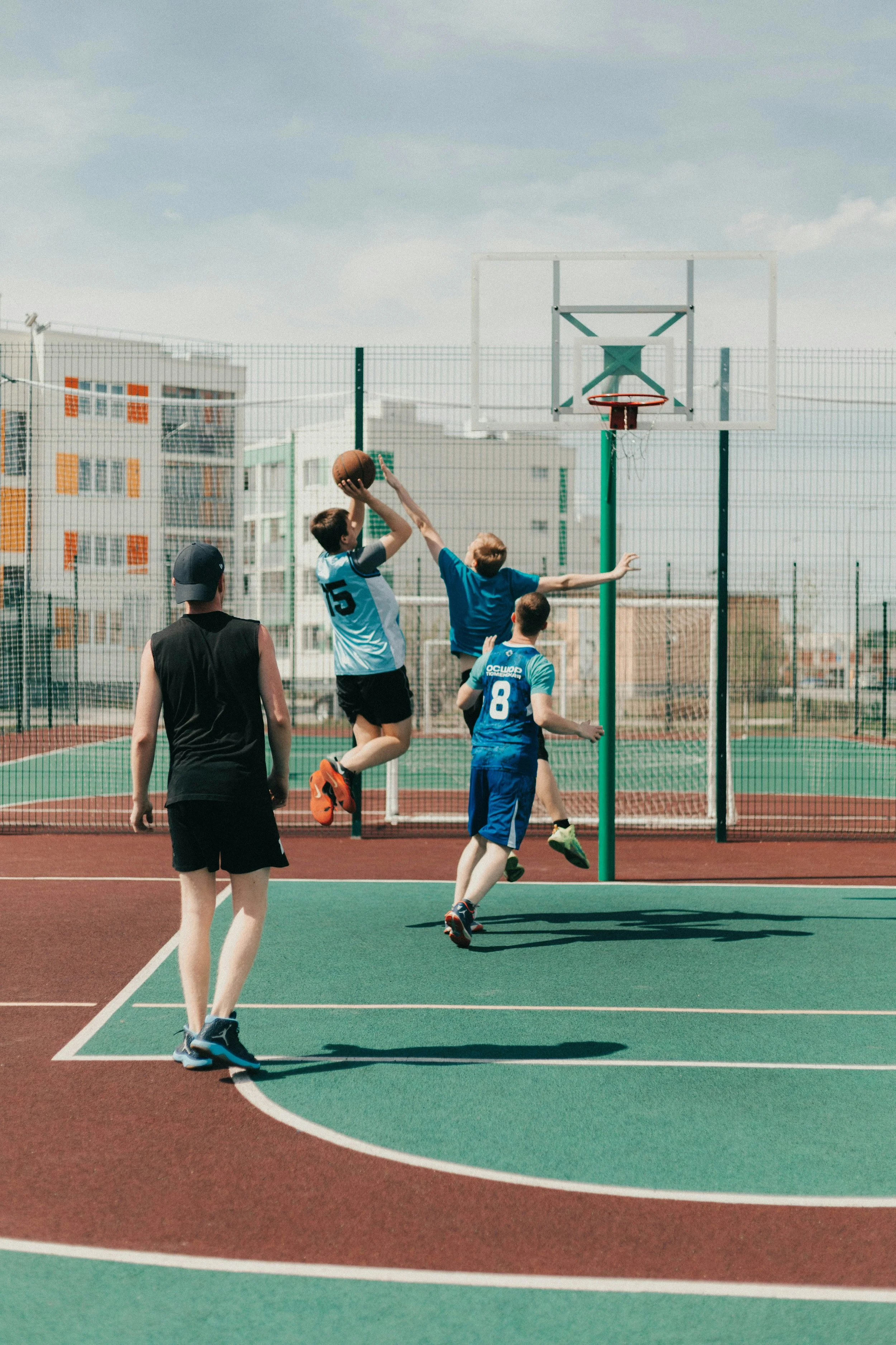 Children playing basketball on an outdoor court surrounded by a fence, with buildings in the background.