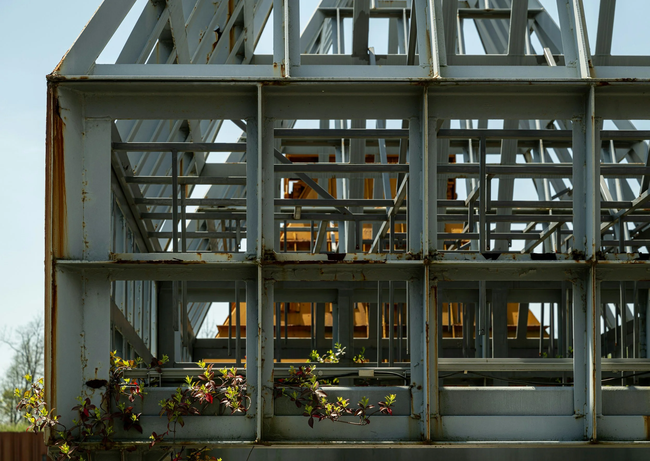 Close-up of a steel structure frame, with visible rust spots and geometric design, against a cloudy sky. Part of a construction or industrial site.