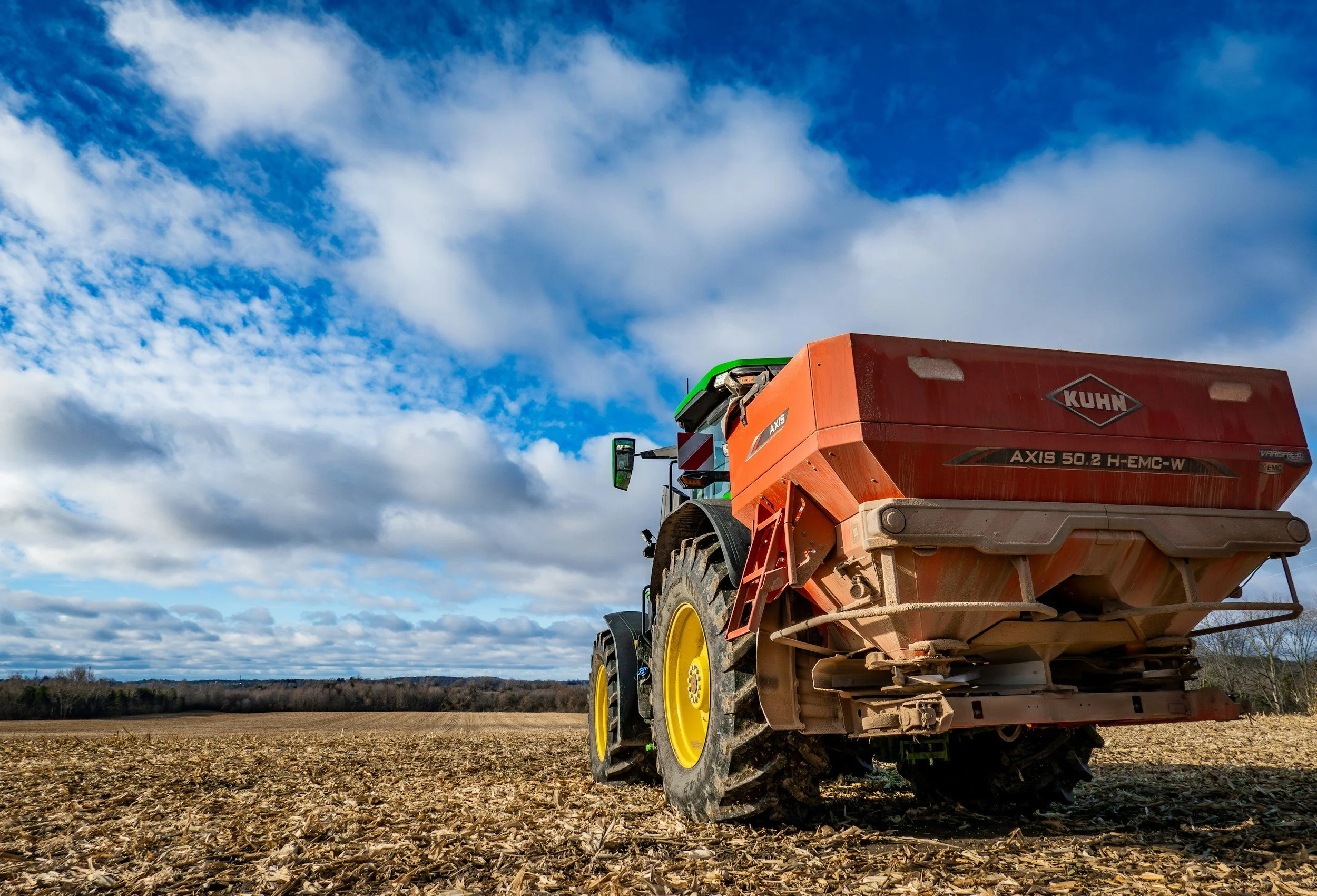 A tractor with a red fertilizer spreader attachment working in a field on a partly cloudy day.