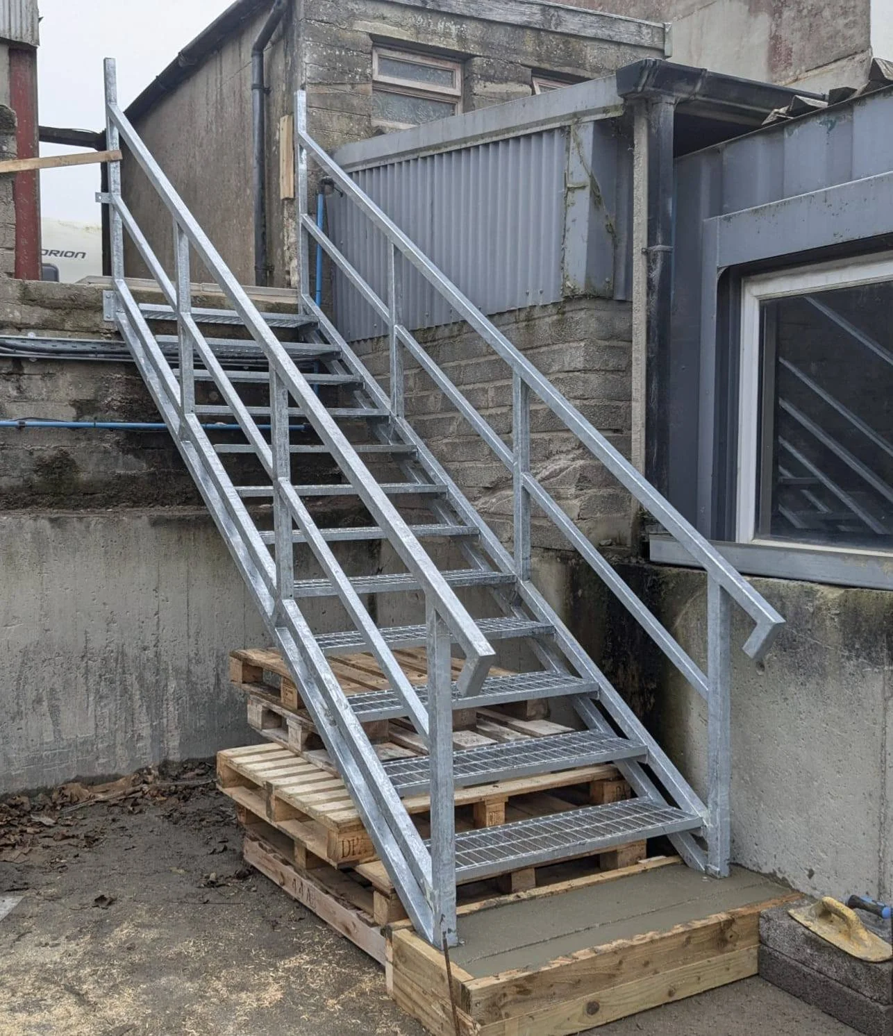 Metal outdoor staircase with handrails leading up to a building, supported on wooden pallets with some concrete at the base, next to a concrete wall and a window.