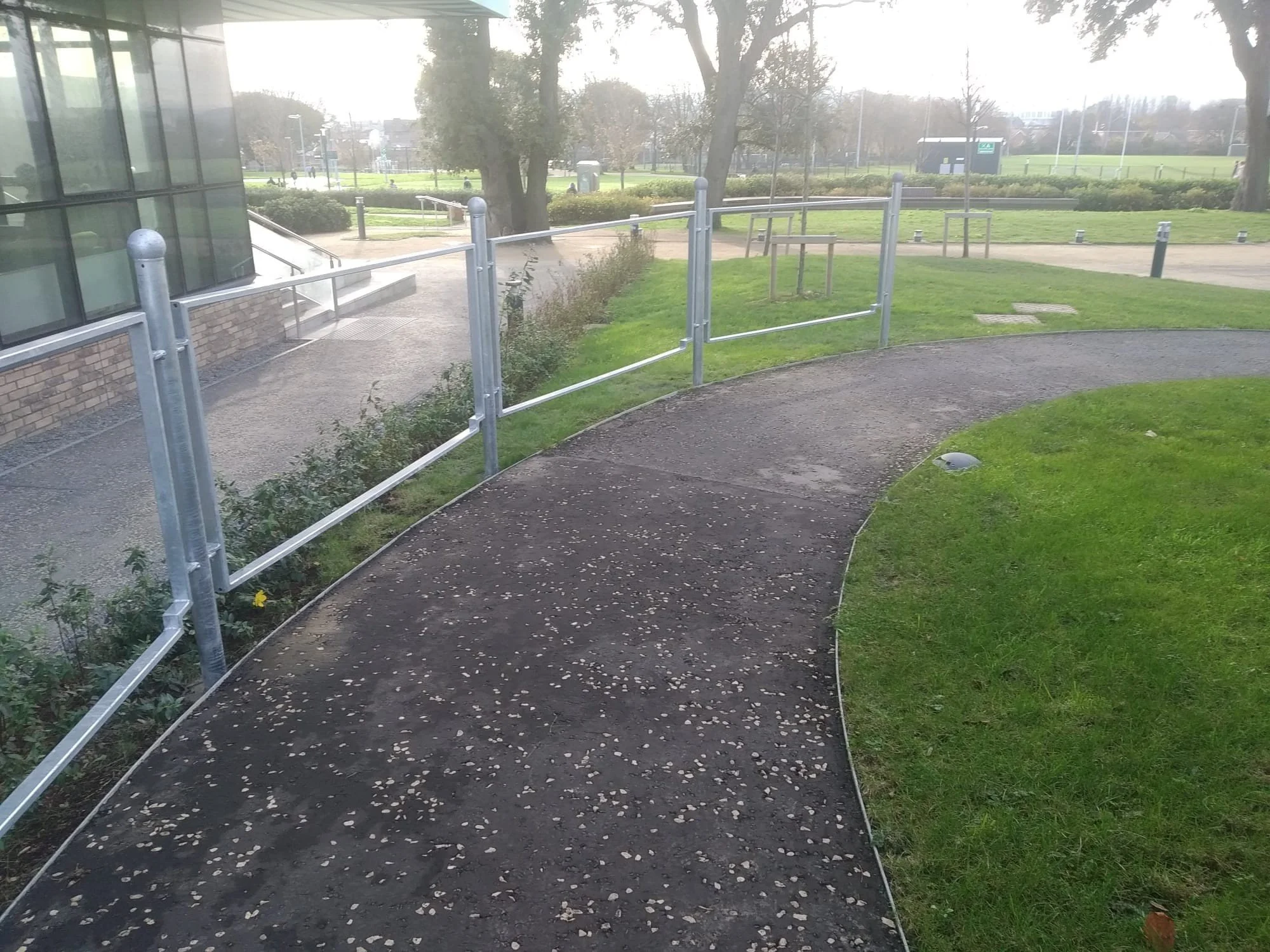 A curved outdoor pathway with a metal railing on one side, adjacent to a grassy area, near a building with large windows, and an open field with trees and a few structures in the background.