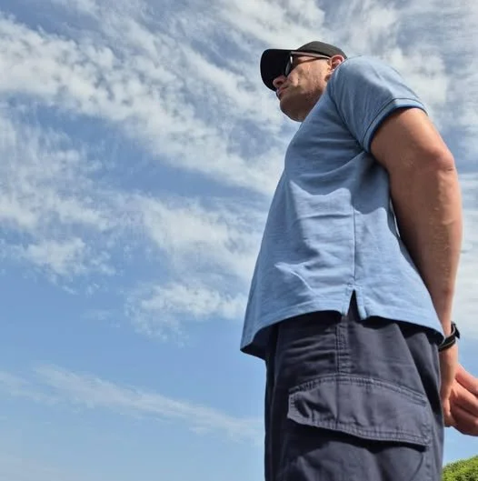 A man in a blue t-shirt, black cap, and sunglasses stands against a blue sky with scattered clouds, facing to the right with his hands behind his back.