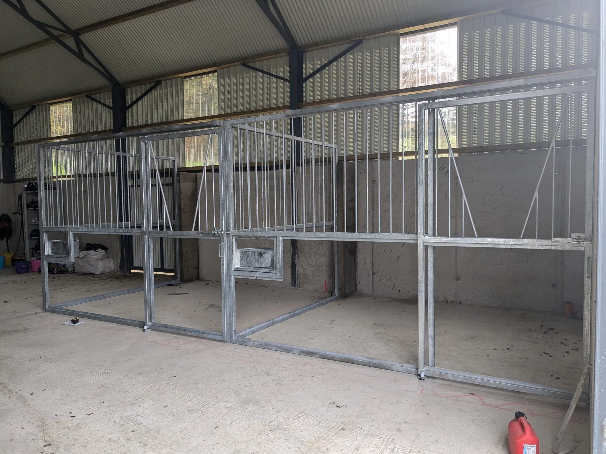 Metal horse stalls under construction inside a barn or riding arena with concrete walls and a metal roof.
