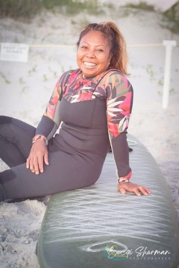 A woman with a bright smile sitting on a paddleboard at the beach, with sandy surroundings and a background of a rocky shoreline.