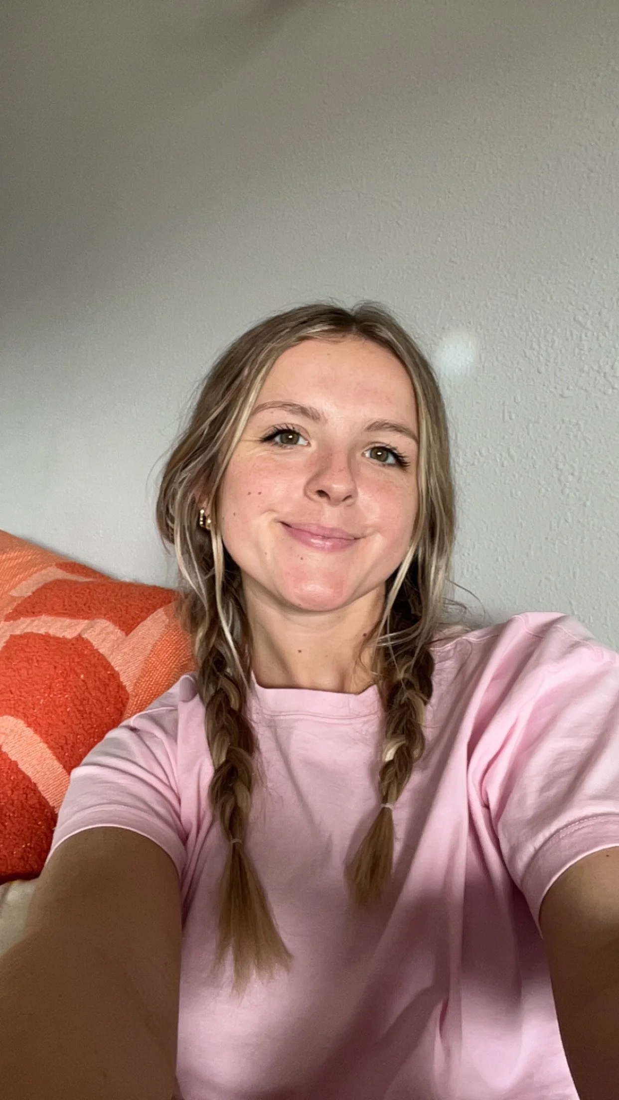 A young woman with light brown hair styled in two braids, wearing a pink t-shirt, smiling and taking a selfie indoors next to an orange patterned cushion.