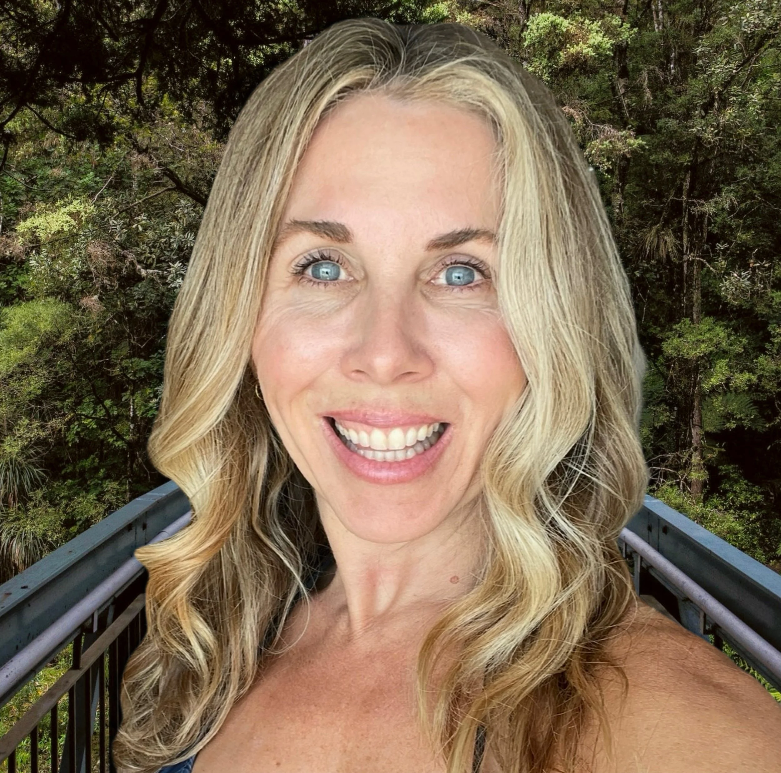 A woman with blonde hair and blue eyes smiling, standing on a balcony with a forest background.