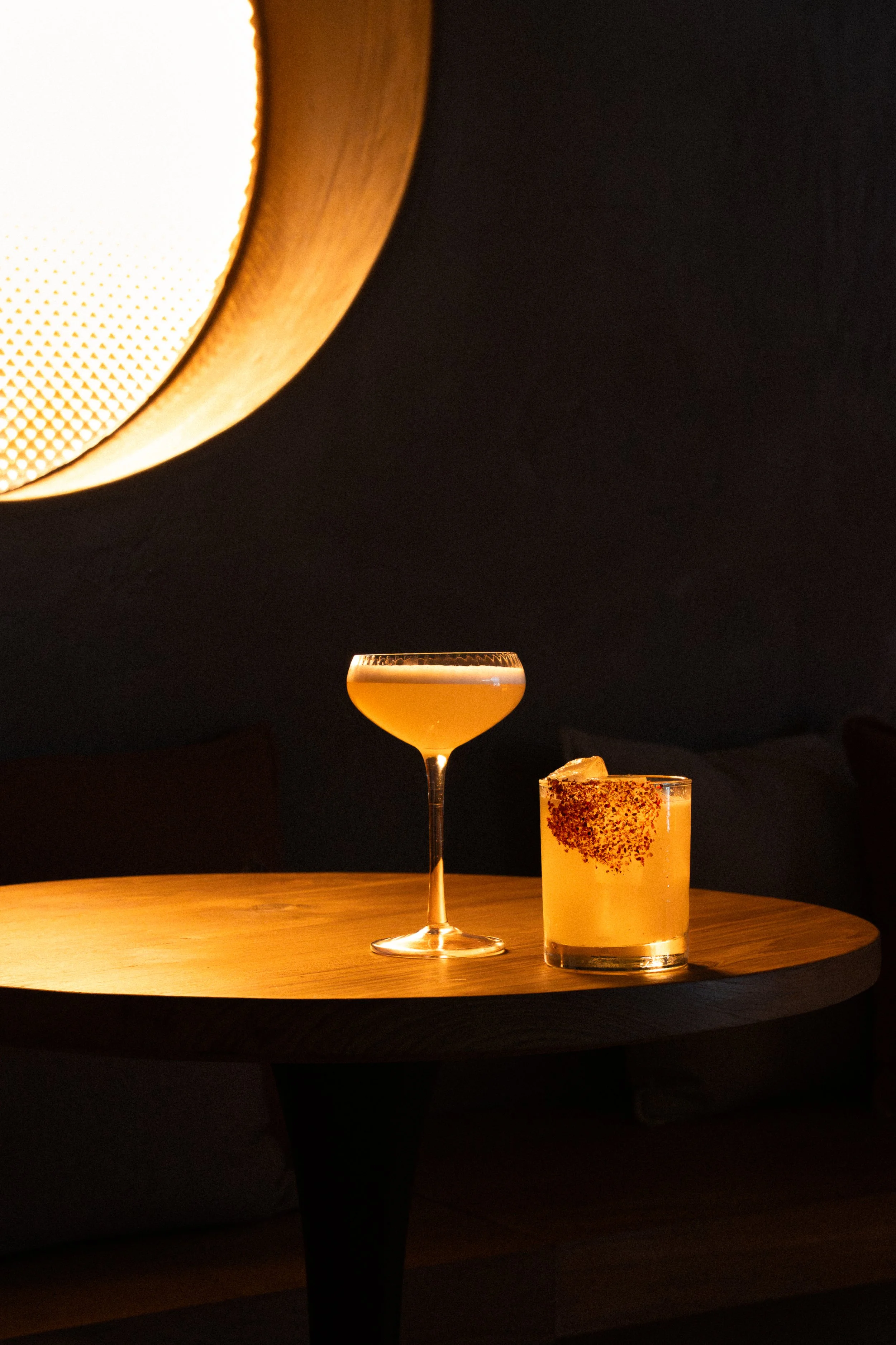 Two cocktails on a round wooden table, one in a coupe glass and the other in a rocks glass, illuminated by a warm ceiling light.