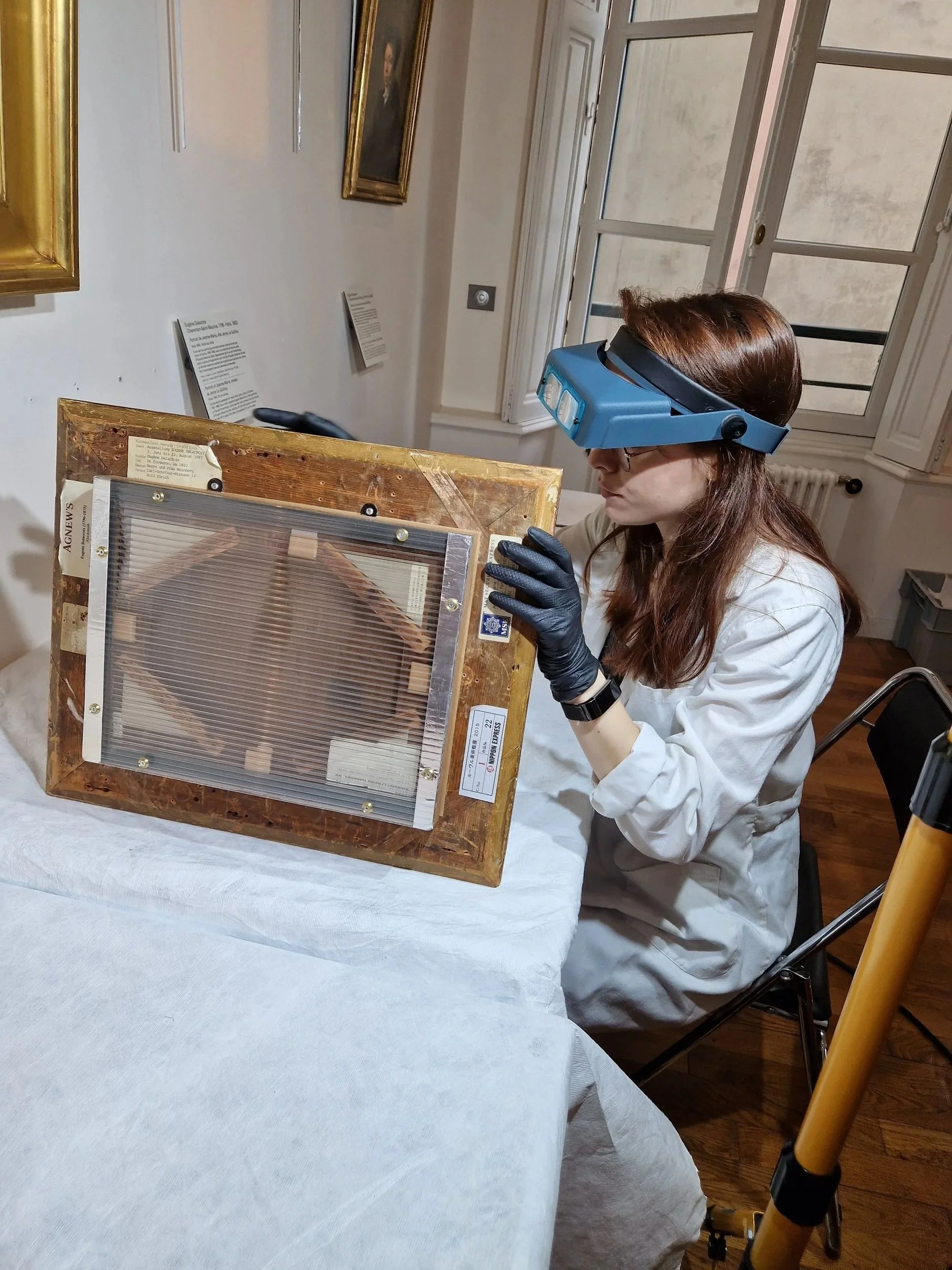 Une femme portant des gants et un lab coat examine un objet mystérieux à l'aide d'une loupe portée sur sa tête, dans une salle d'exposition ou un laboratoire.