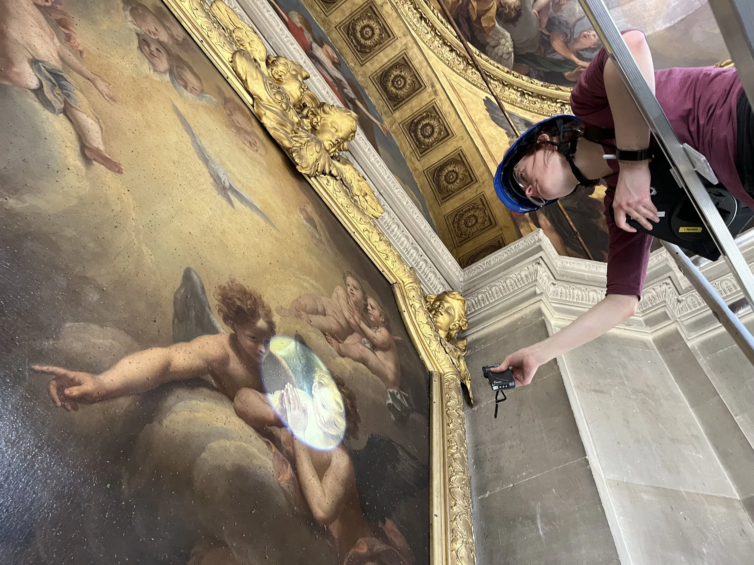 Une personne portant un casque bleu et tenant un appareil photo observe une grande peinture ancienne dans une salle ornée de moulures dorées et de décorations architecturales.