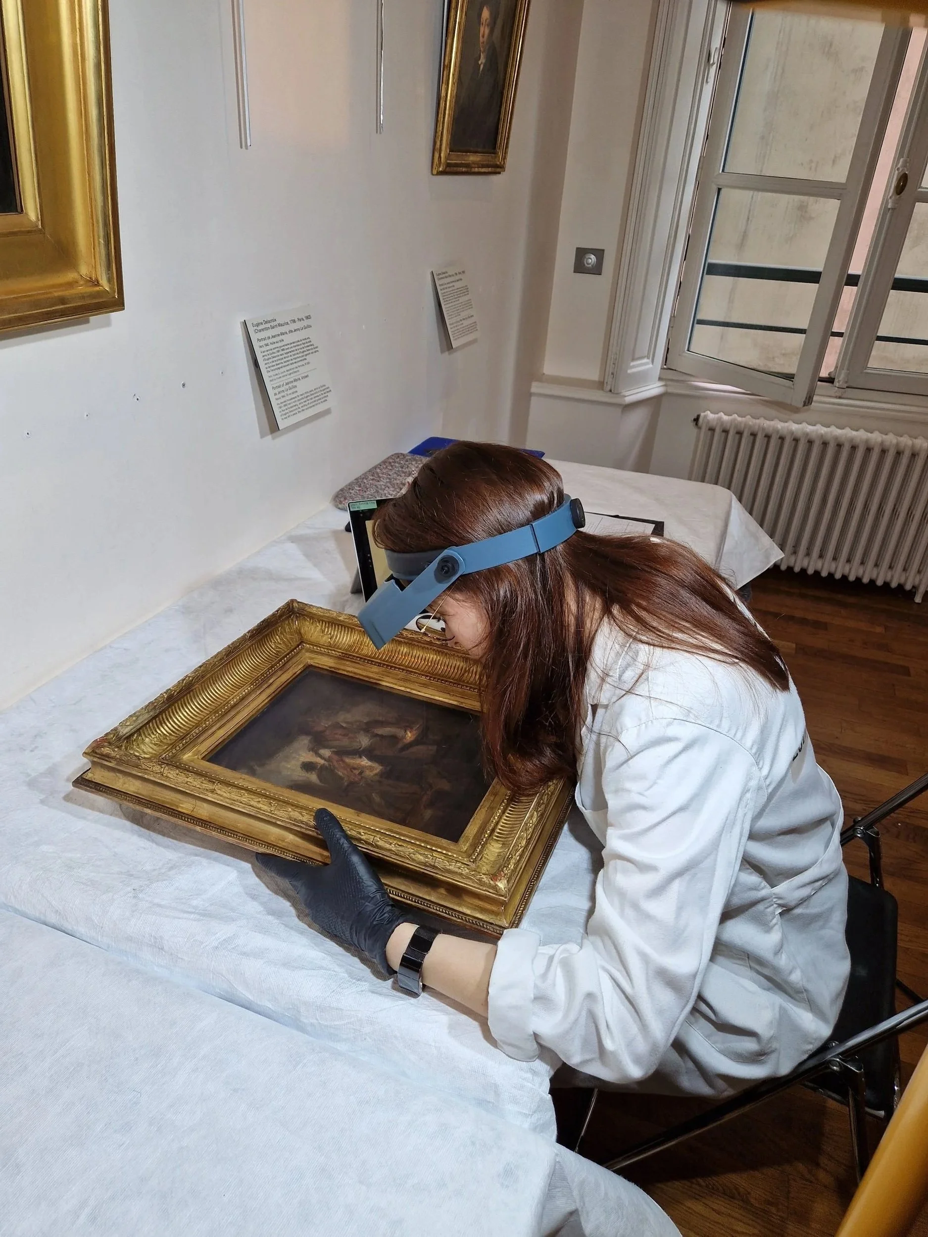 Une conservatrice examine une peinture ancienne dans une galerie d'art, utilisant une loupe et portant une visière de protection.
