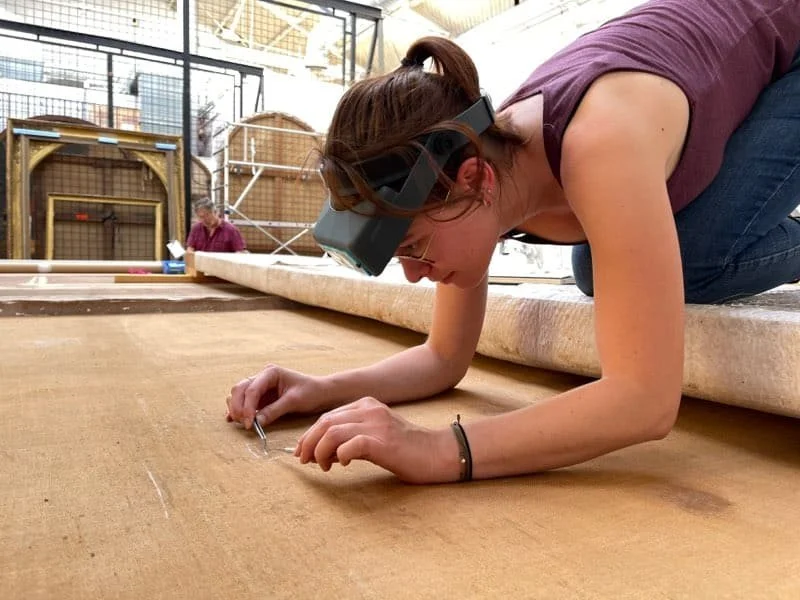 Une femme porteuse de lunettes de protection travaille en position accroupie à même le sol sur un tapis ancien dans un atelier ou un musée, avec des structures en arrière-plan.