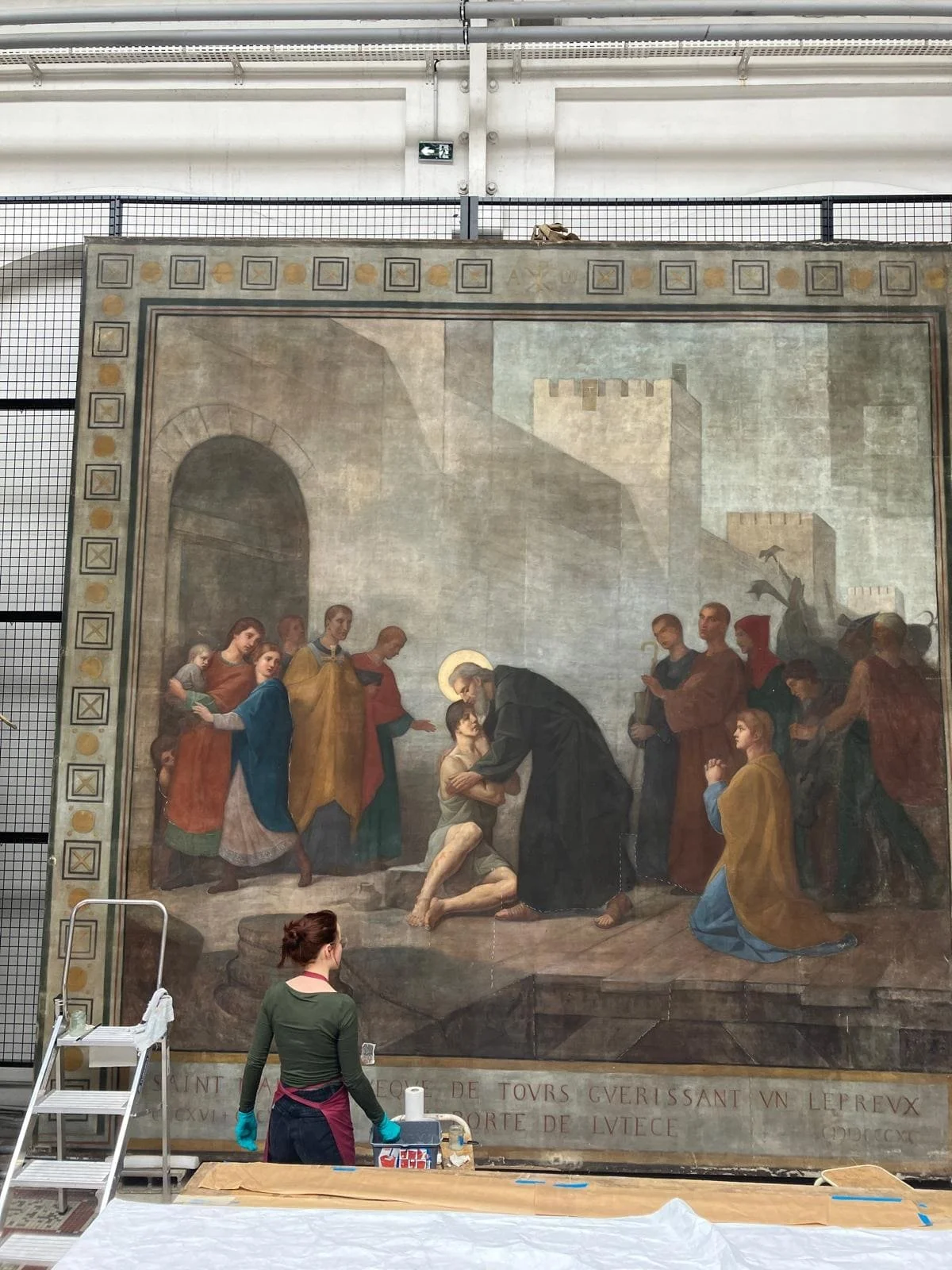 Une femme travaille à la restauration d'une grande peinture religieuse dans un musée ou une galerie, portant des gants bleus.