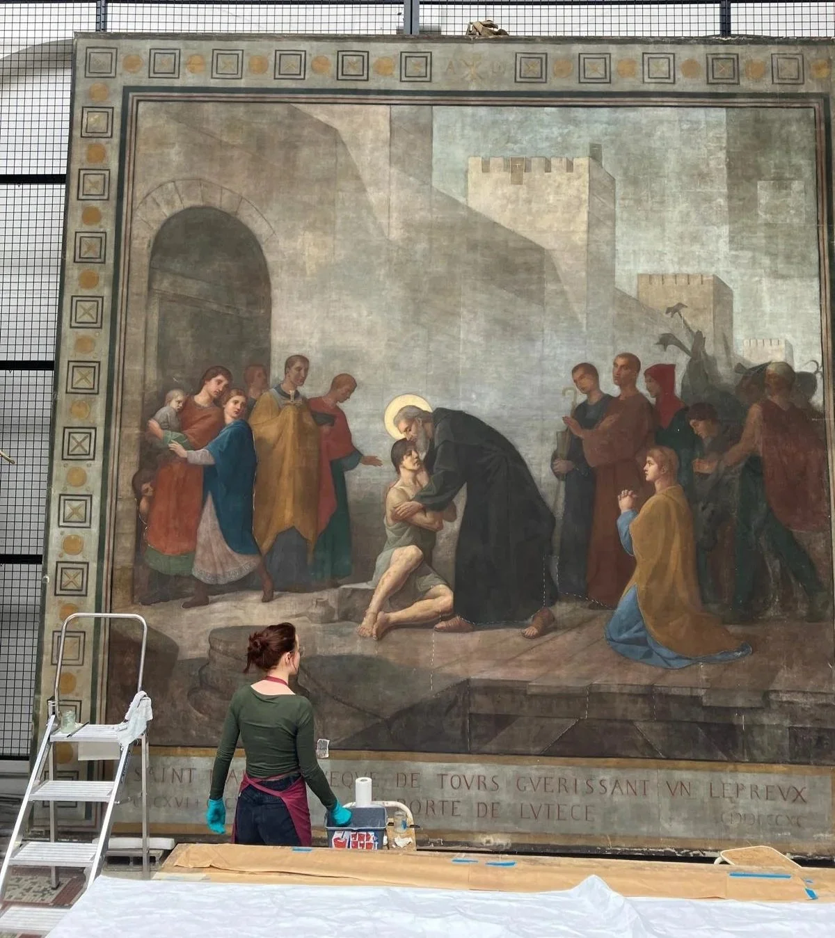 Une femme travaillant sur la restauration d'une grande peinture religieuse représentant la purification d'un homme par un religieux, avec plusieurs autres figures en arrière-plan.