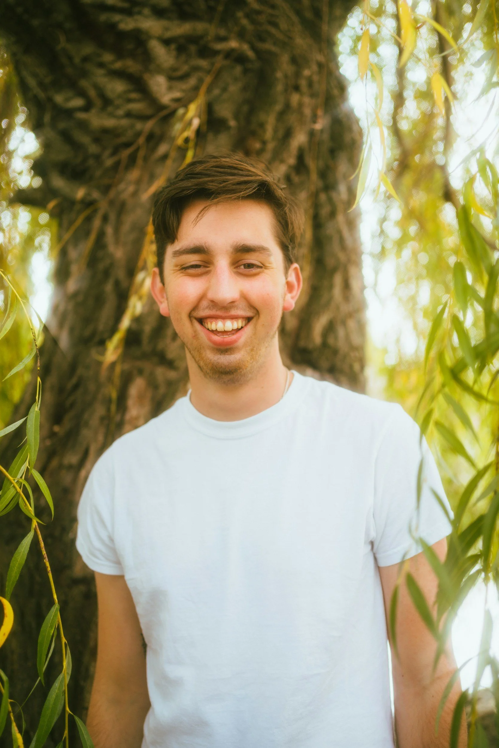 A young man in a white t-shirt smiling outdoors with a large tree and green leaves in the background.