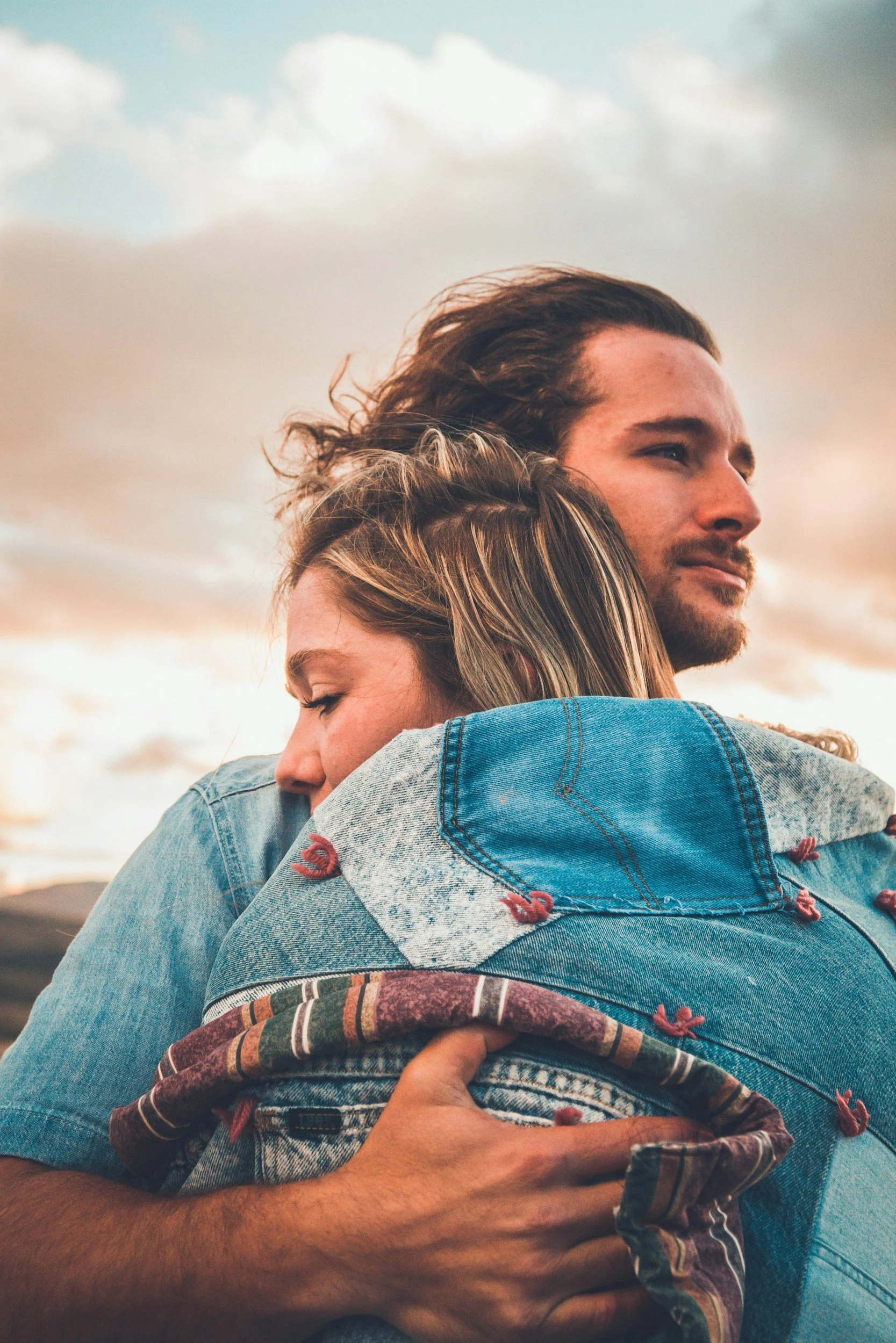 A couple hugging outdoors during sunset, with the woman resting her head on the man's shoulder and both appearing content.