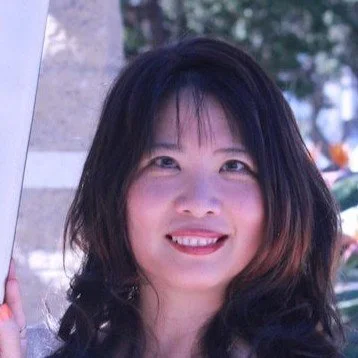 A woman with dark hair smiling outdoors, with trees and plants in the background.