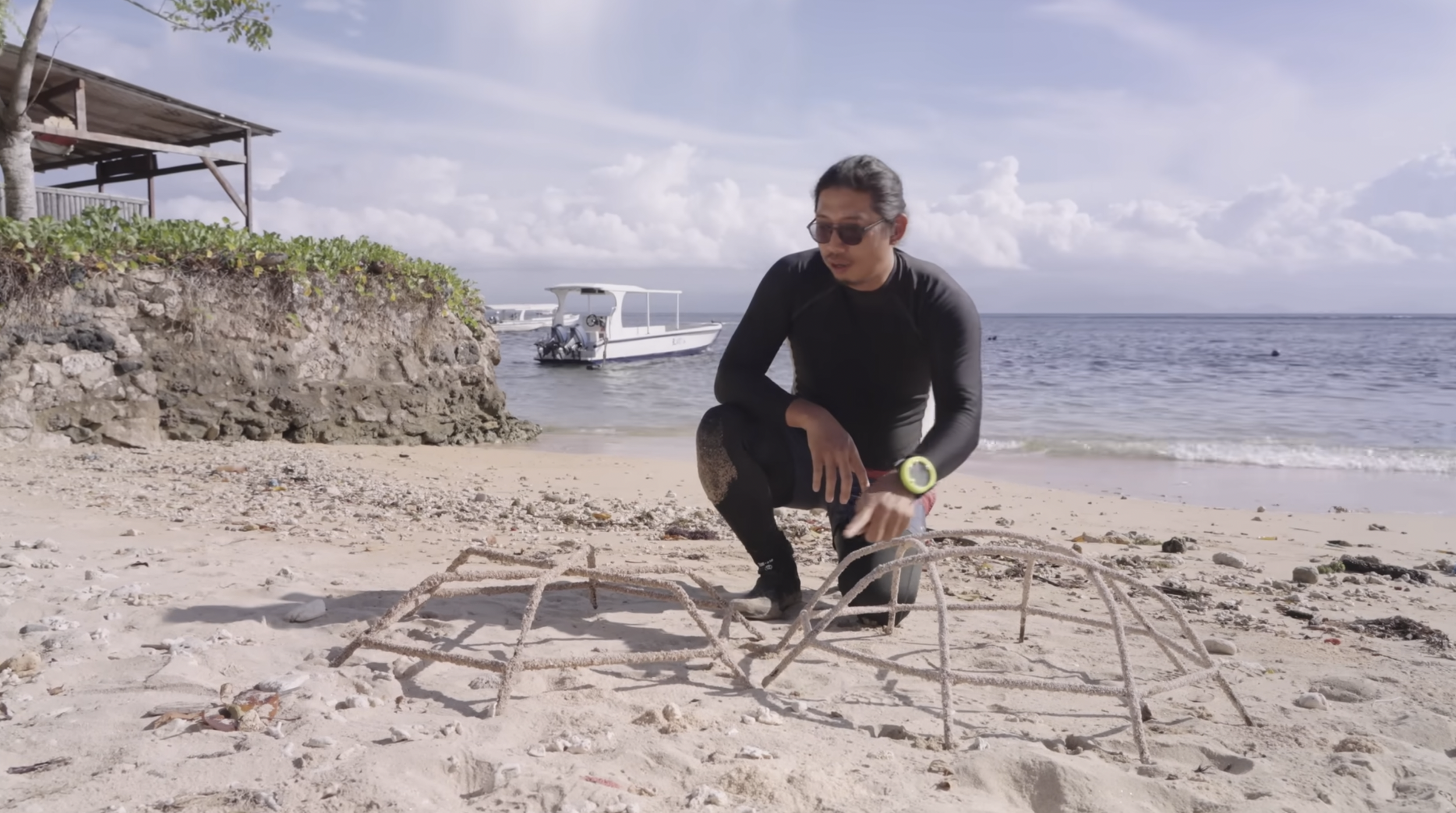 Man wearing black rash guard and sunglasses at beach, assembling a large metal crab sculpture, with boat in water and cloudy sky in background.