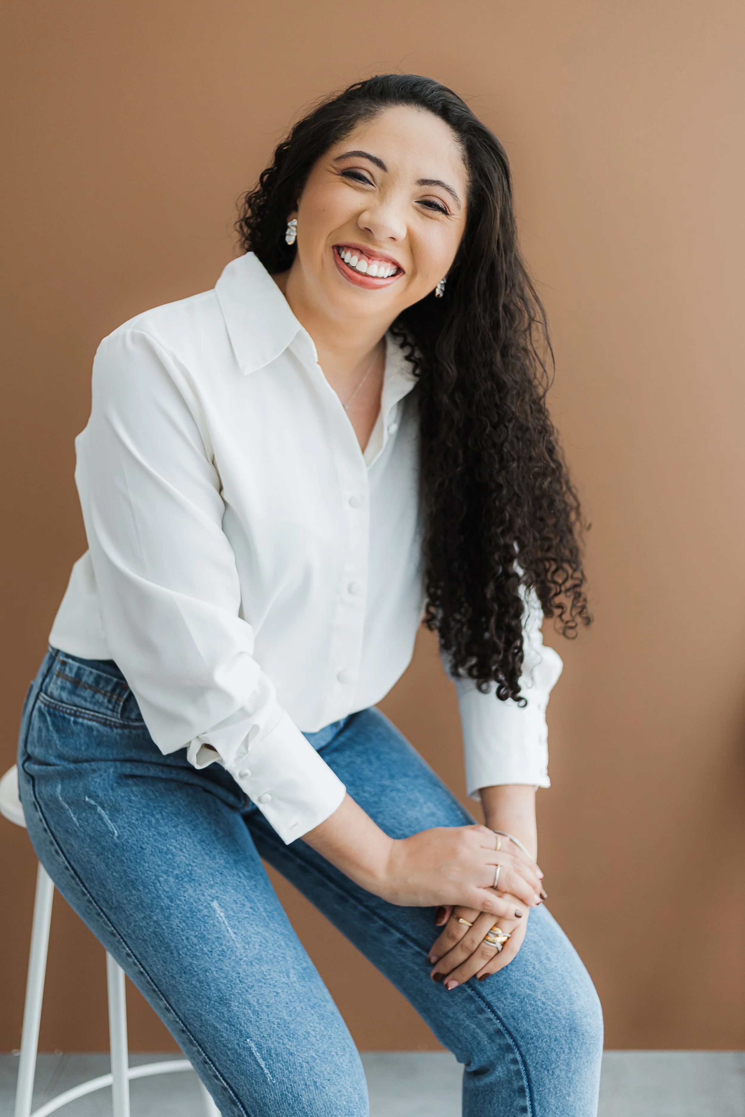 A woman with long curly dark hair, smiling and winking, wearing a white button-up shirt and blue jeans, sitting against a plain beige wall.