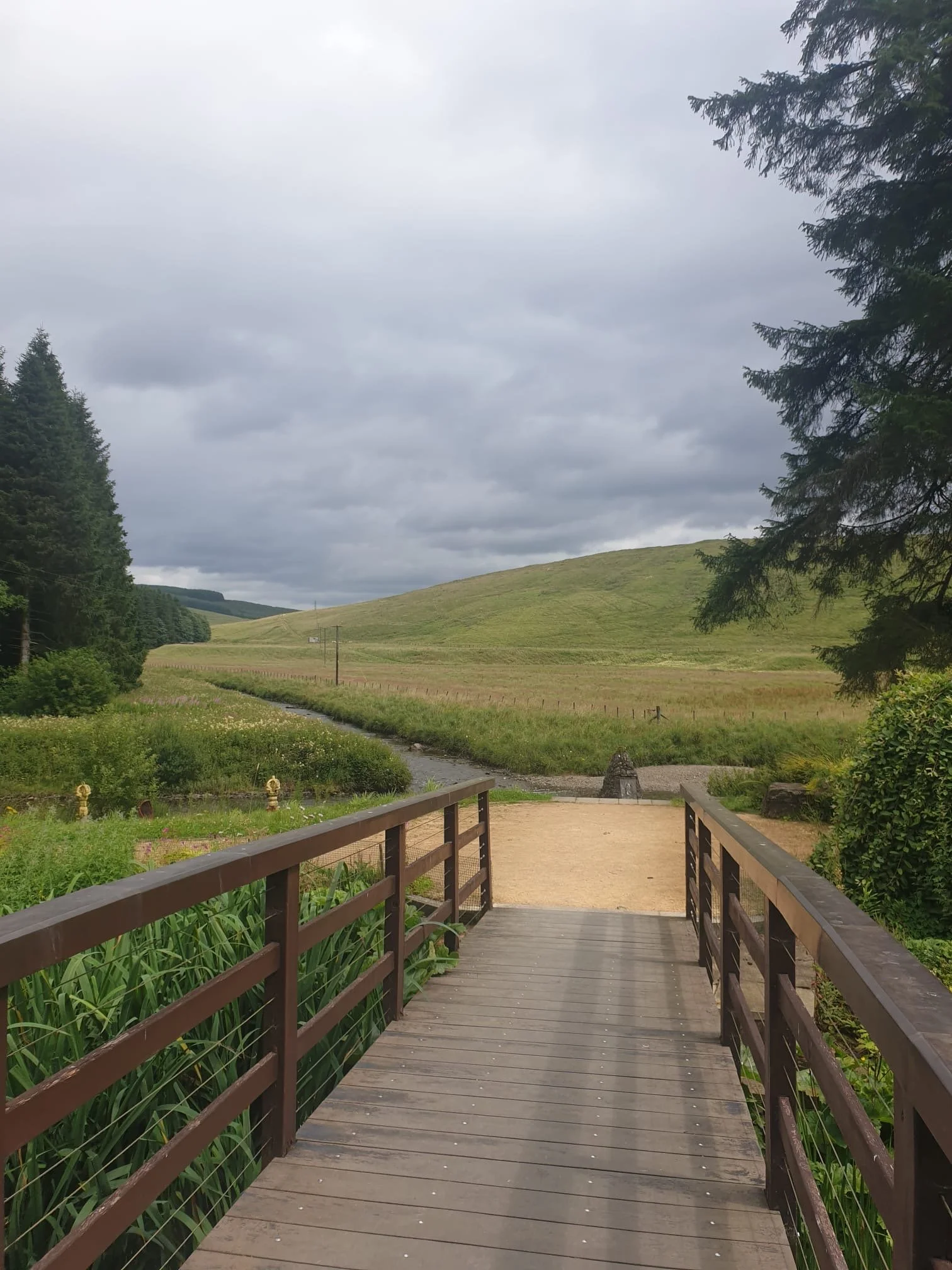 View of a wooden bridge leading to a grassy field with rolling hills and tall trees on the left and right, under a cloudy sky.