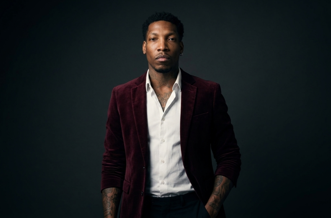 Portrait of a man with tattoos wearing a white shirt and burgundy velvet blazer against a dark background.