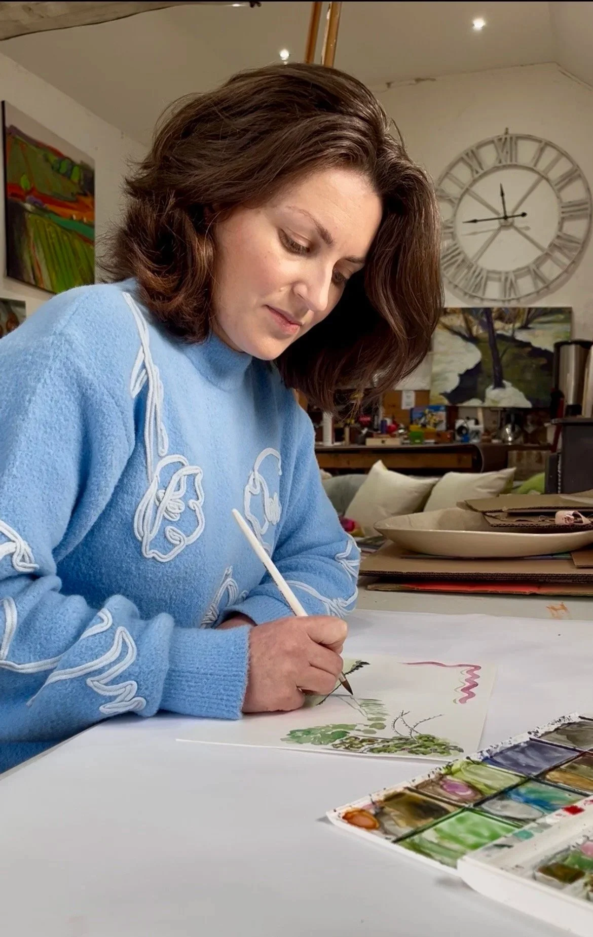 A woman with brown hair wearing a light blue sweater, painting with a fine brush on paper at a table with watercolor paints.