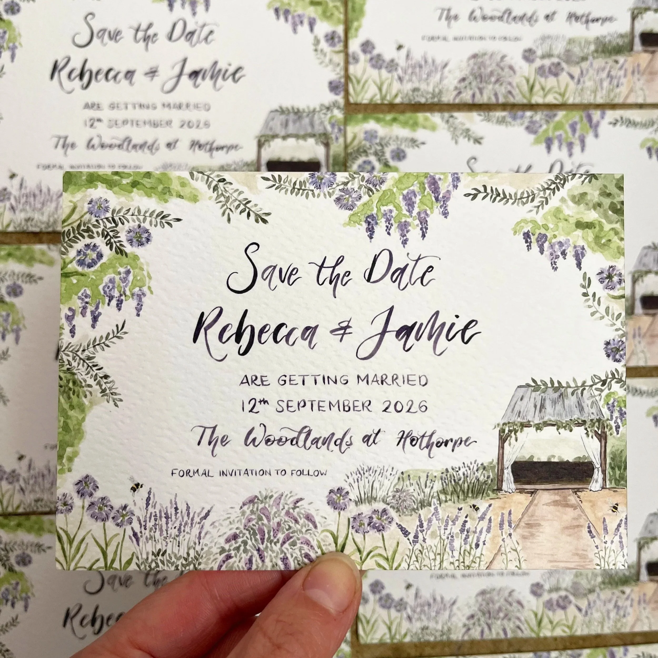A hand holding a botanical-themed wedding save the date card with lavender, greenery, and a small wooden outdoor gazebo illustration.