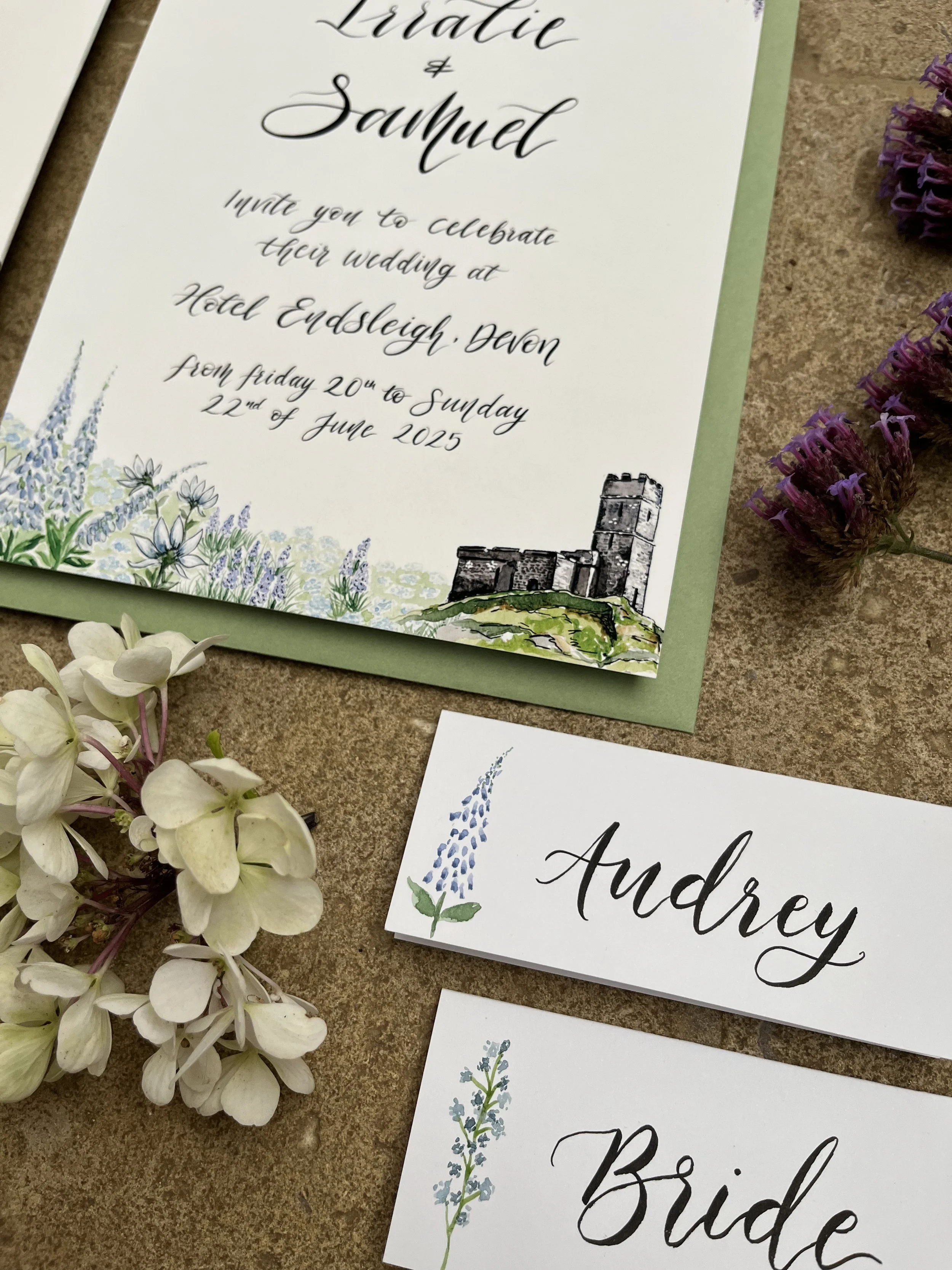 Watercolor painting of a two-story brick house with a steep gray roof, surrounded by green bushes and trees, and a flower bed in the foreground.