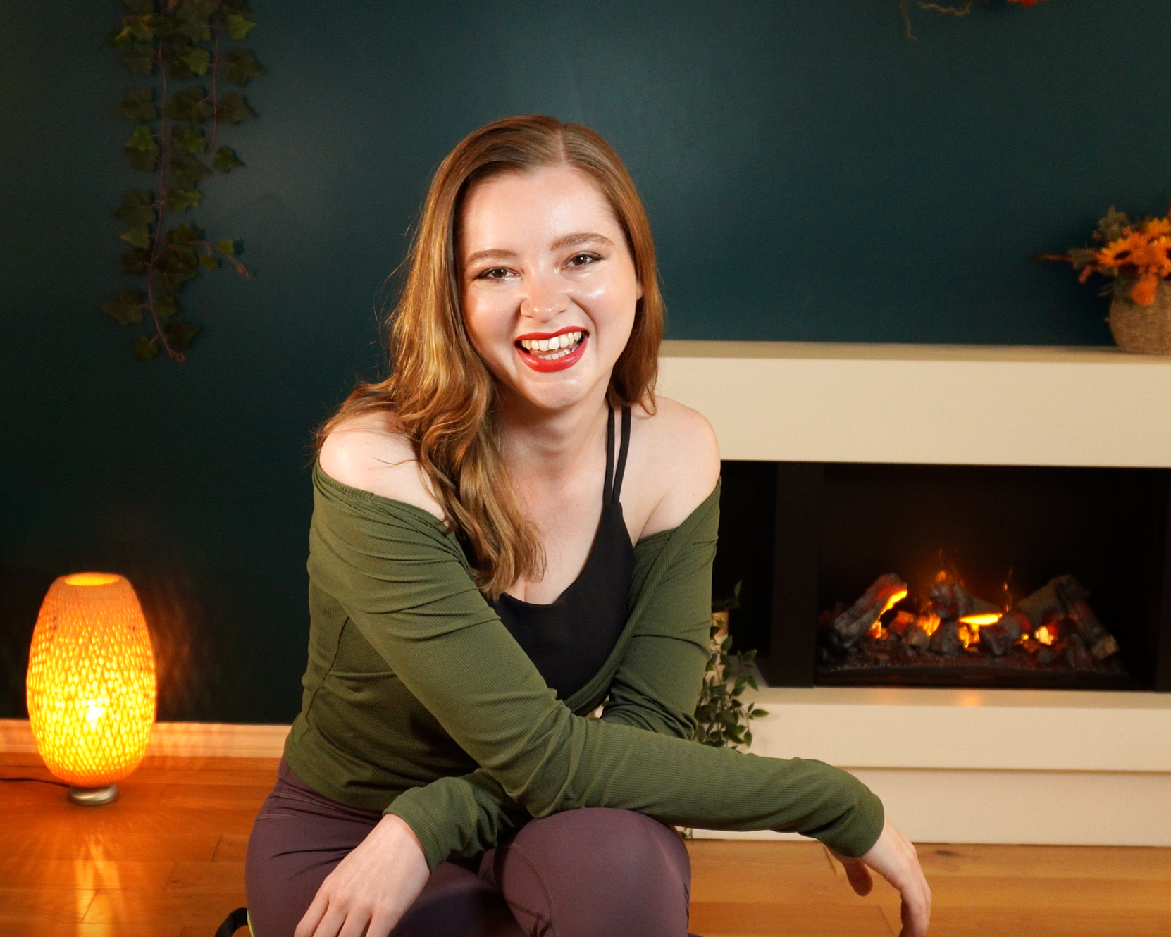 Jenn | Pilates teacher smiling at camera. She has red hair, red lipstick and is sitting in front of a fireplace