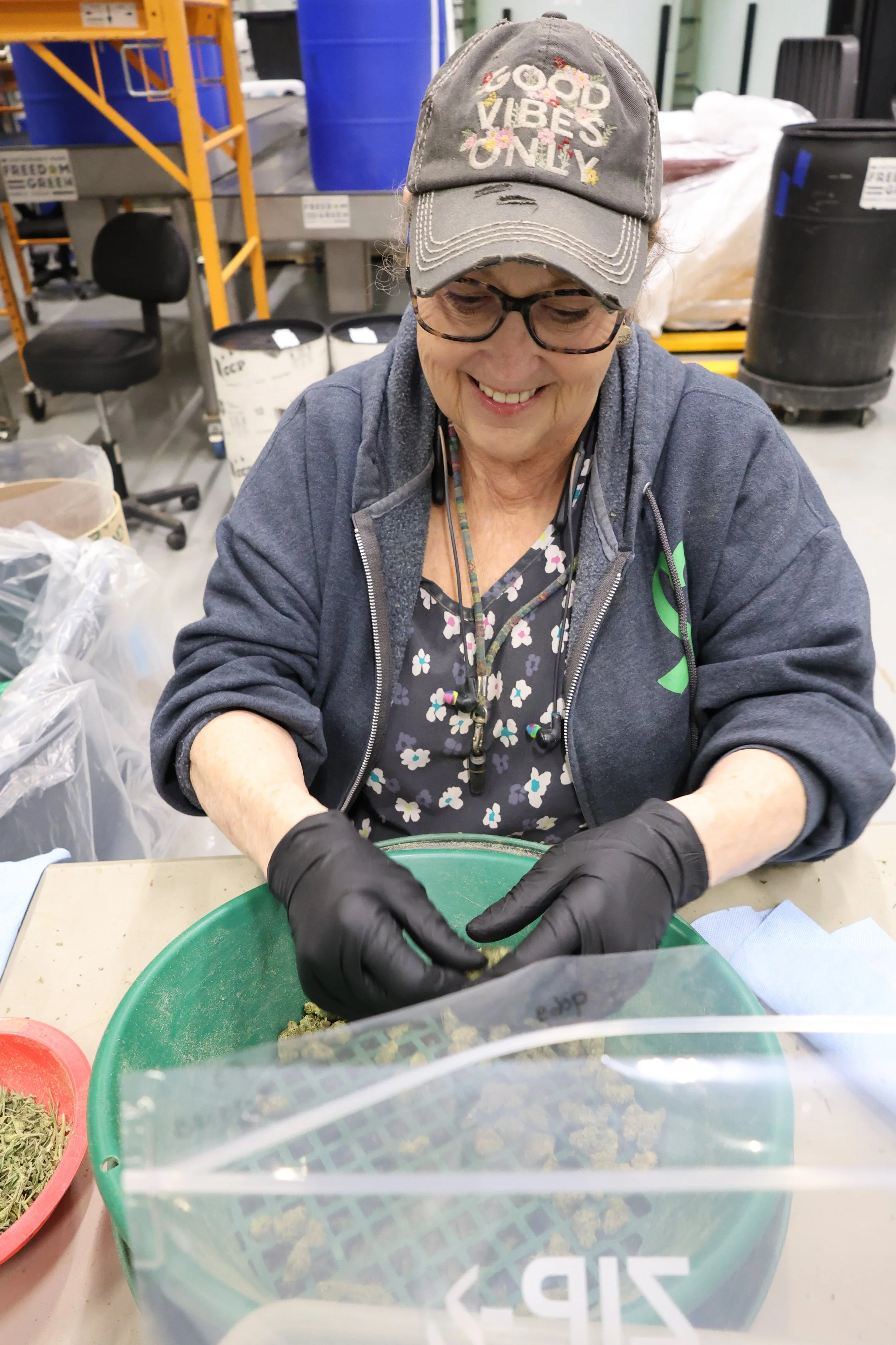 A woman wearing a cap with the words 'Good Vibes Only,' glasses, a floral shirt, a gray hoodie, and black gloves is working with cannabis buds in a large green container in an indoor facility.