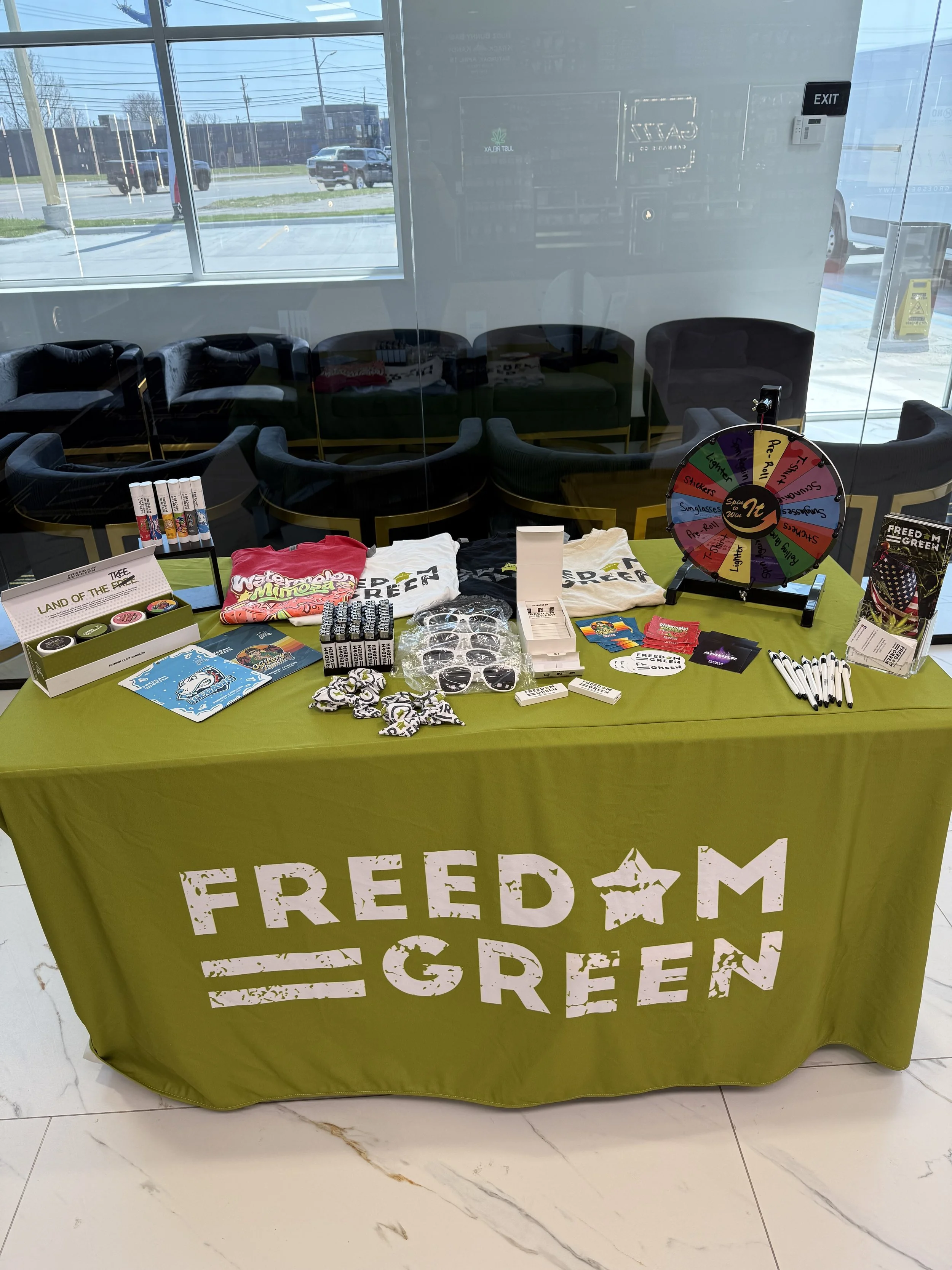 Table covered with green cloth displaying promotional items and merchandise for Freedom Green, with the organization's name and logo, inside a building near large windows and waiting chairs outside.
