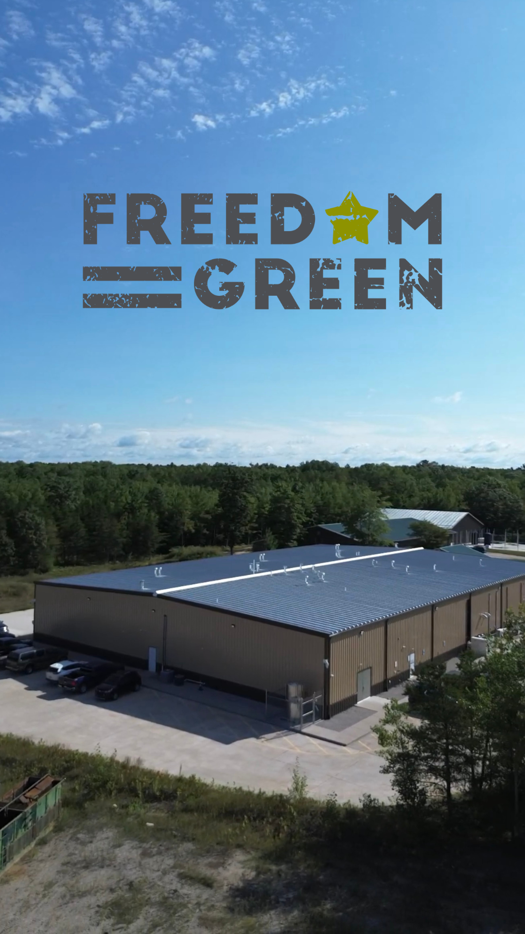 A large industrial building with a metal roof, surrounded by parking lot, with dense forest in the background and a blue sky overhead.