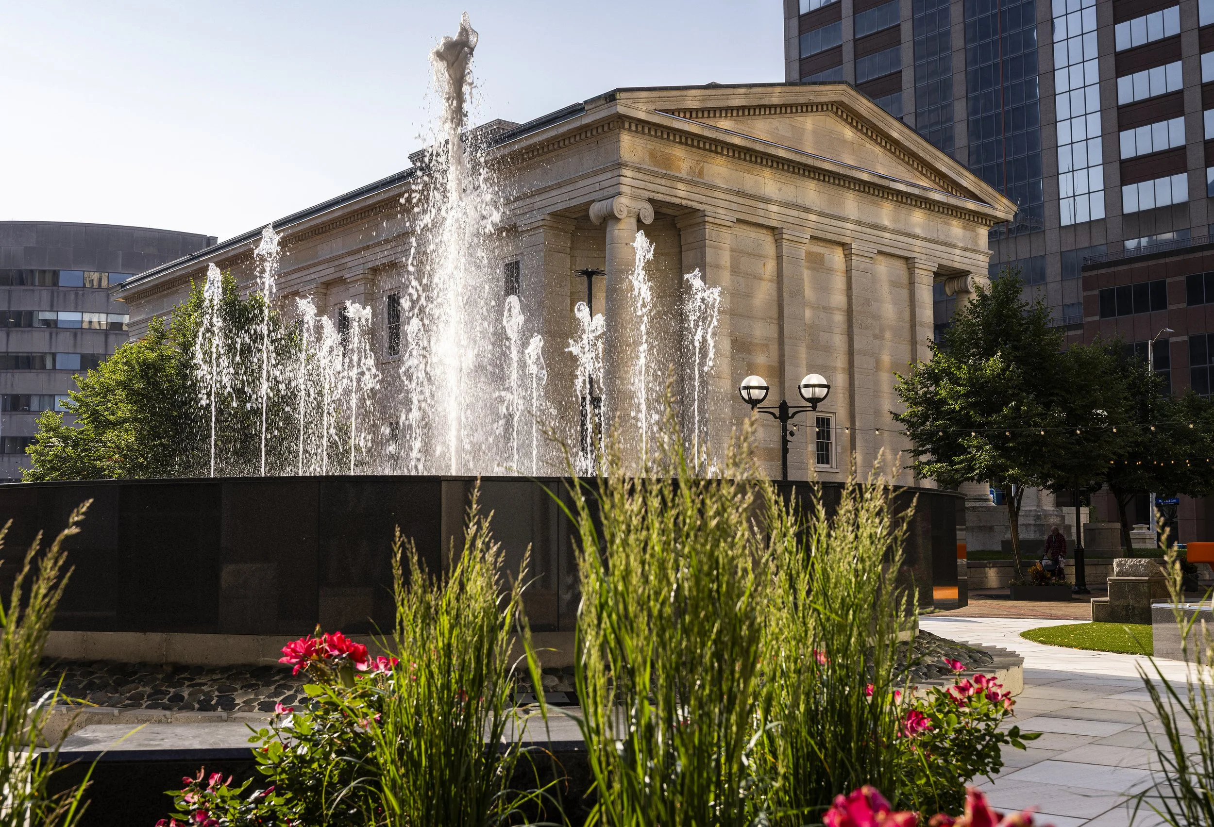 Historic stone building in Dayton, Oh with large columns, fountain with water jets, surrounding trees, and flower garden in an urban park area.