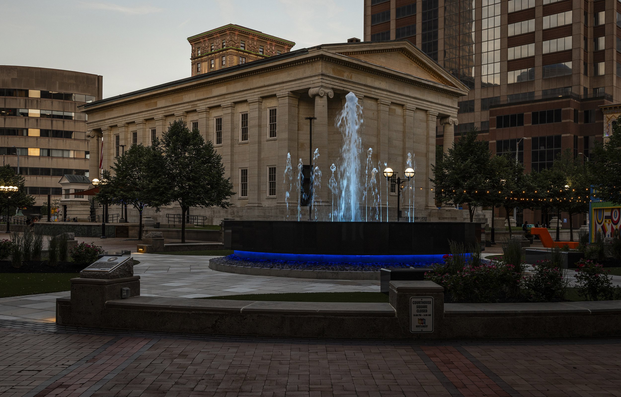 Downtown Dayton, OH, fountain illuminated in blue water, surrounded by trees and benches, in front of a historic building with columns and modern high-rise buildings in the background.