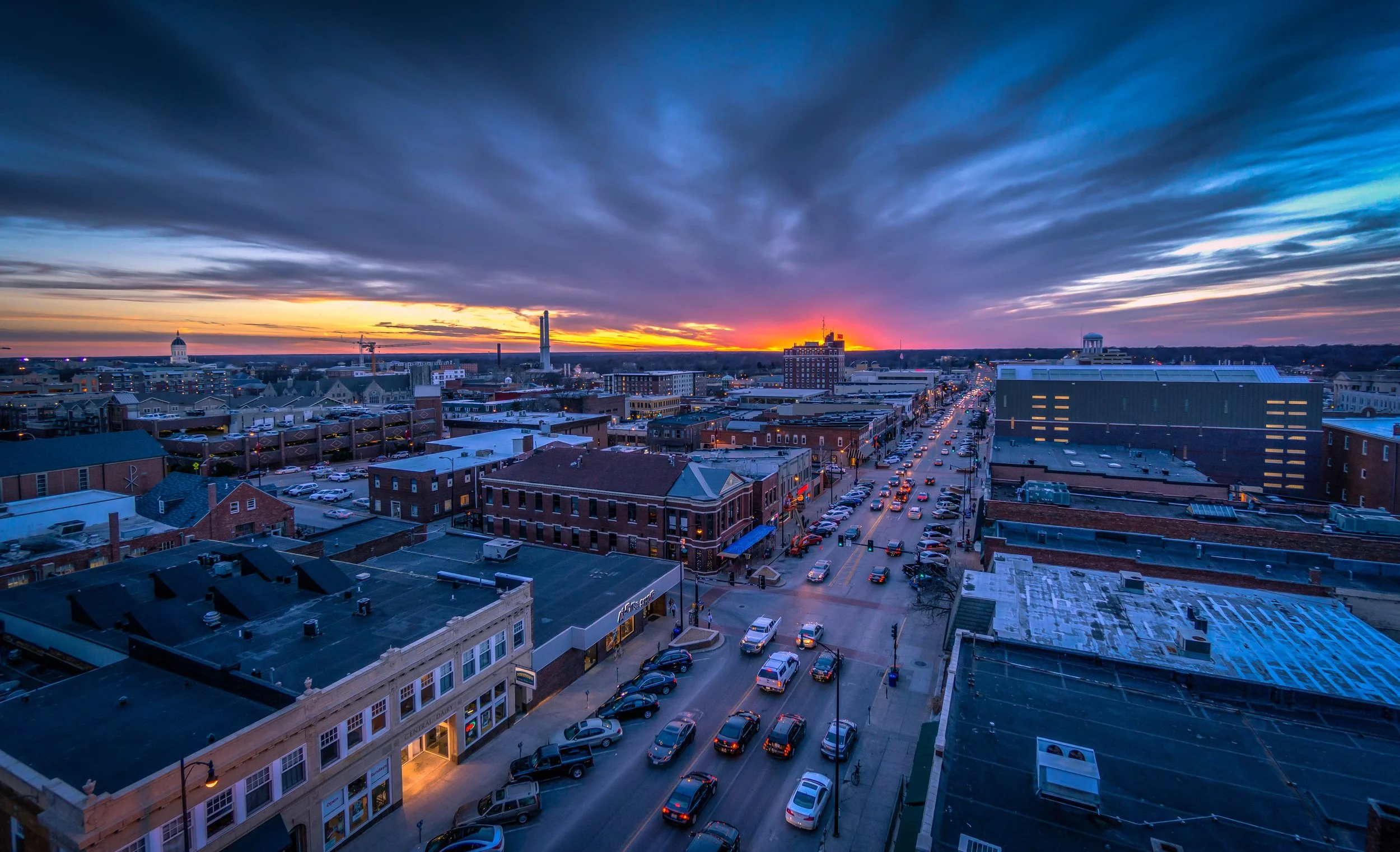 A city street during sunset with cars, buildings, and a colorful sky with clouds.