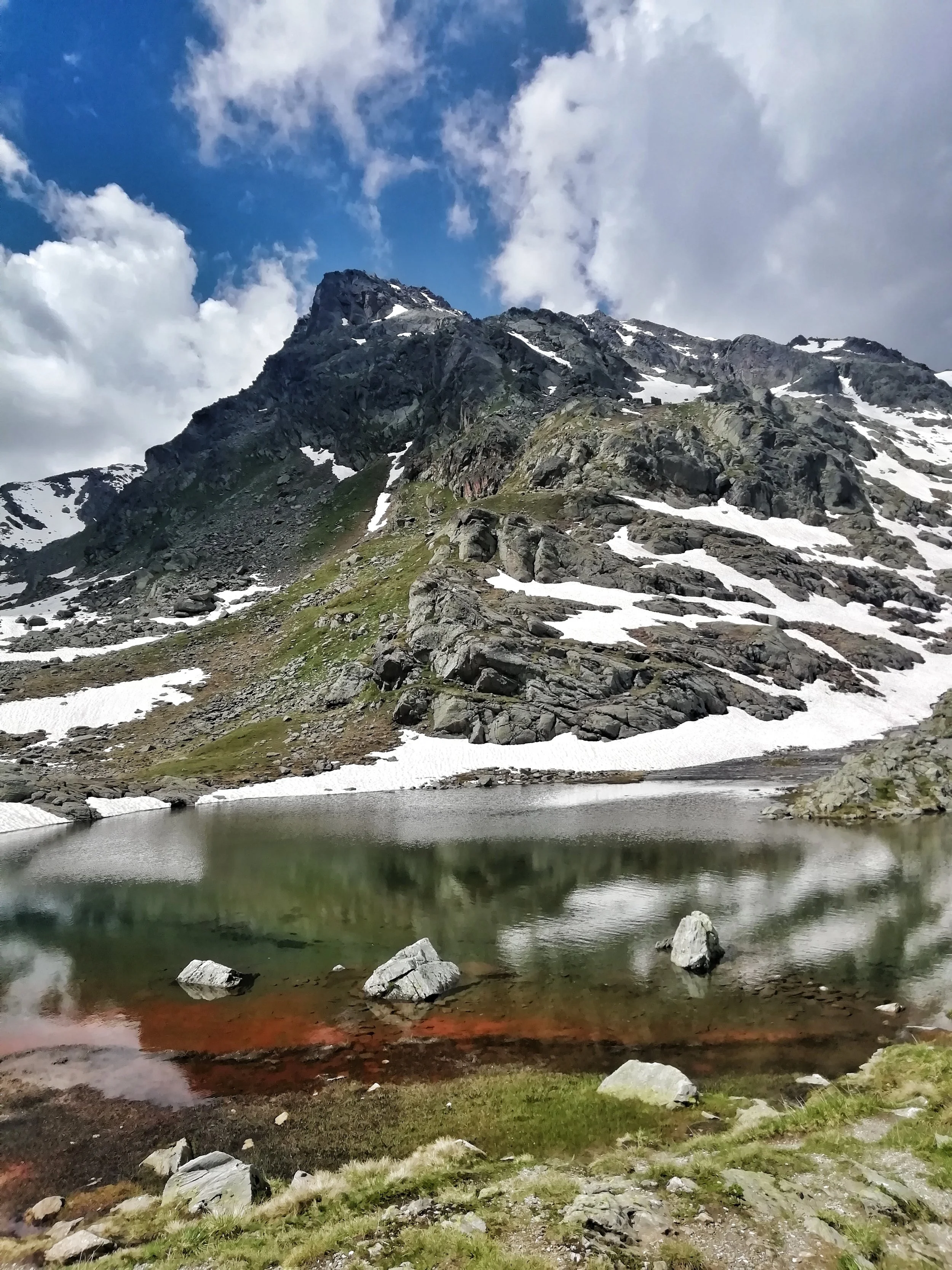 Berglandschaft mit einem glatten See im Vordergrund, umgeben von Felsen und einem schneebedeckten Hang, mit einer hohen Bergspitze im Hintergrund. Skandinavische Gebirgsregion bei bewölktem Himmel.