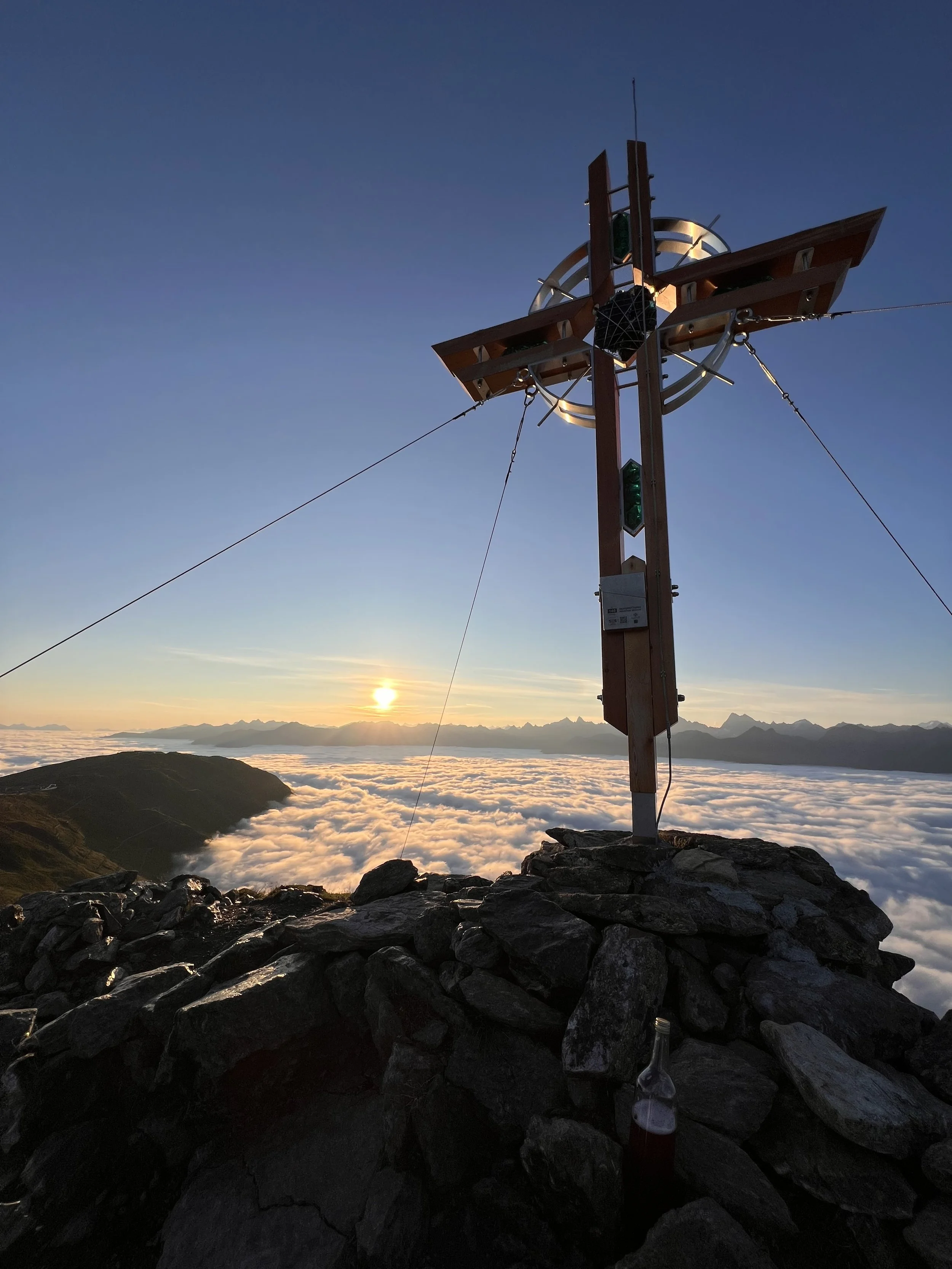 Berggipfel mit Holzkreuz bei Sonnenaufgang, Wolken über Tälern, in der Ferne entfernt, mit einer Flasche auf Steinen im Vordergrund.