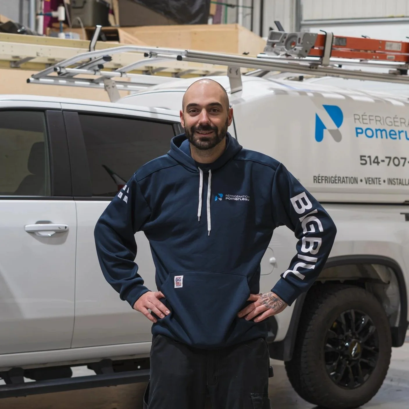 Un homme souriant avec une barbe, portant un sweat à capuche bleu avec le logo 'Réfrigération Pomerleau', se tenant devant une camionnette blanche avec le même logo et des outils de travail sur le toit, dans un atelier de réparation.