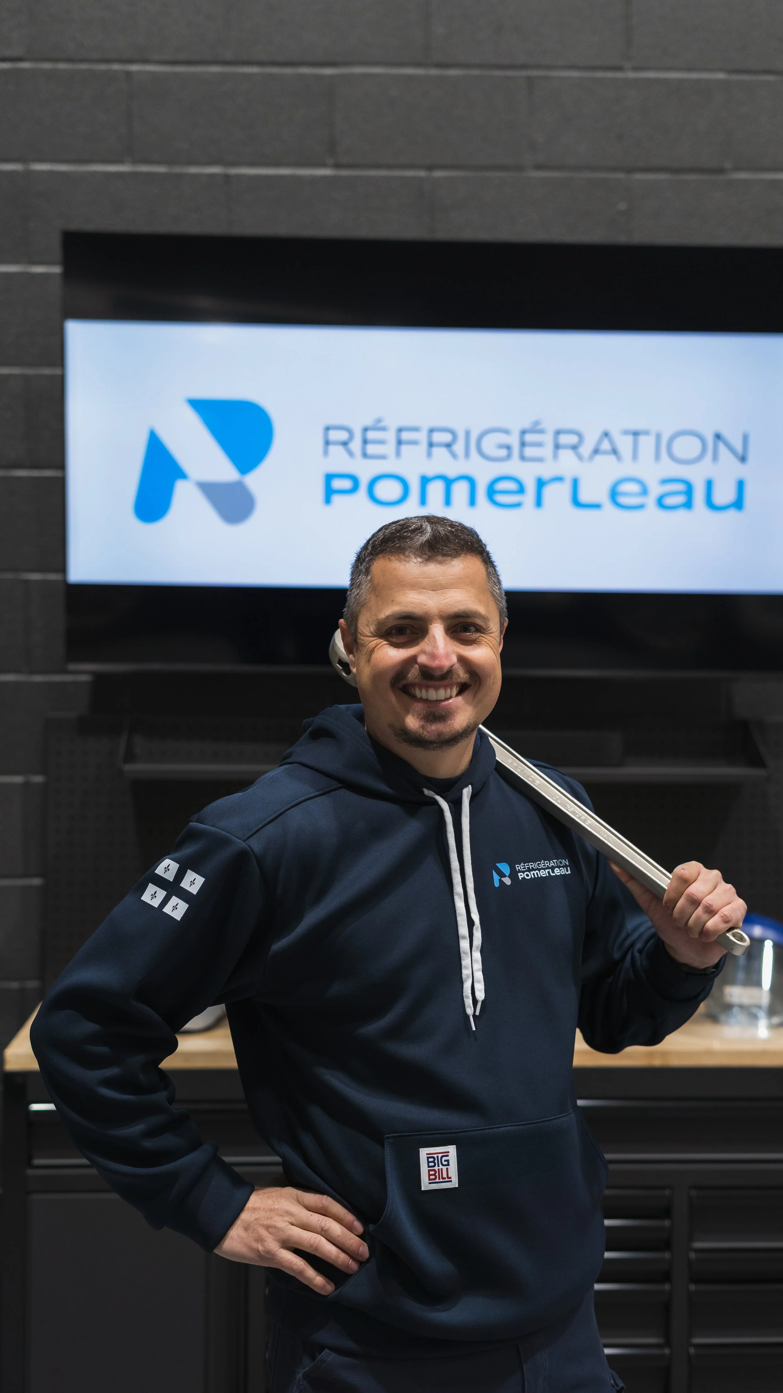 Un homme souriant porte un sweat à capuche bleu foncé avec le logo de Réfrigération Pomerleau, tenant une clé à molette sur son épaule, devant un écran affichant le logo de Réfrigération Pomerleau.