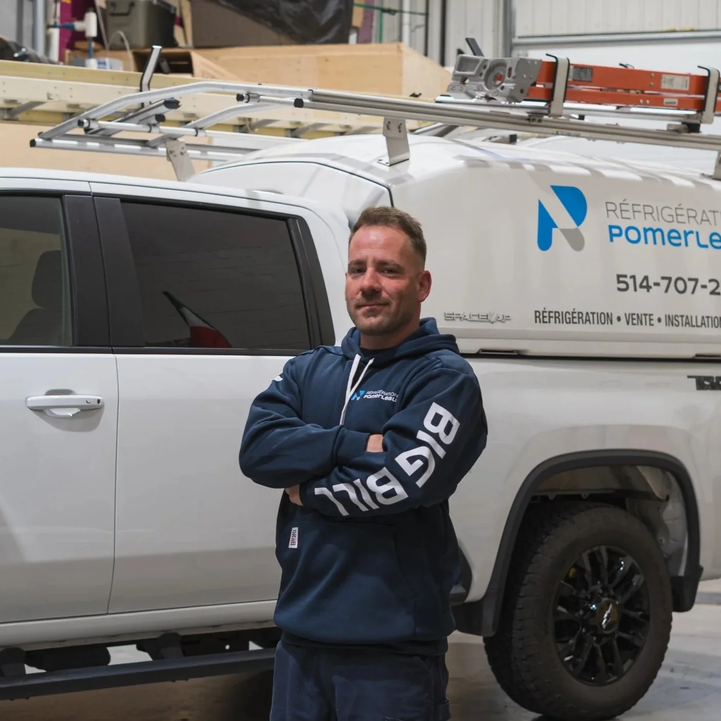Homme posant devant un camion de travaux, portant une veste à capuche bleue avec un logo de la société de réfrigération Pomerleau. Le camion est blanc avec des logos et informations de contact.