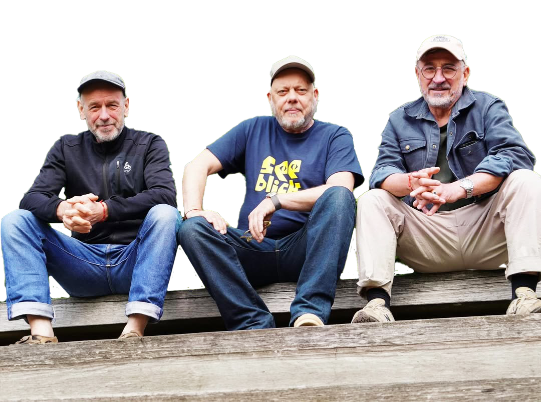 Drei ältere Männer sitzen auf einem Holzsteg, entspannen und schauen in die Kamera, tragen lässige Kleidung und Mützen.